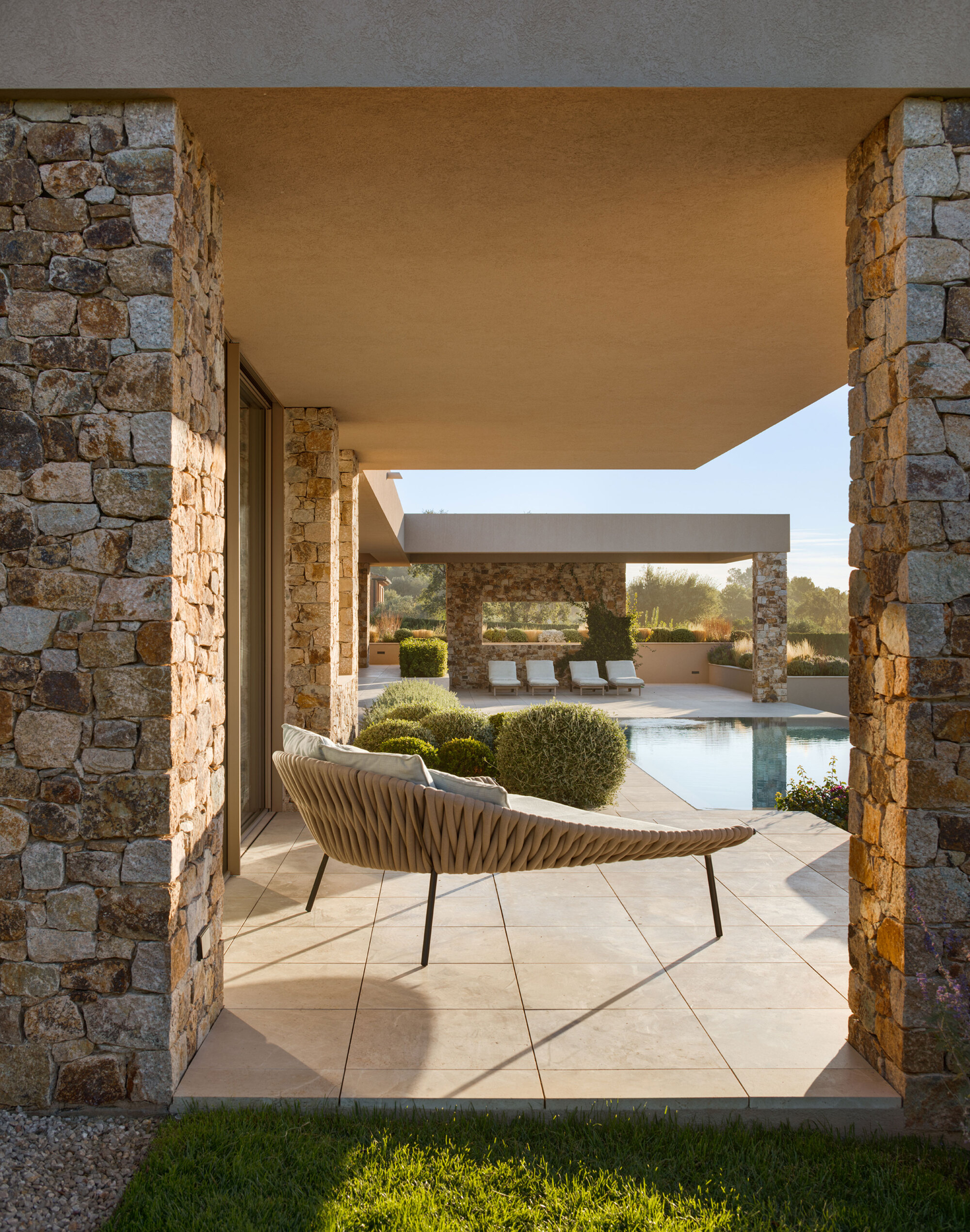 Woven daybed on a paved porch between stone columns, looking across a pool with sun loungers in morning light