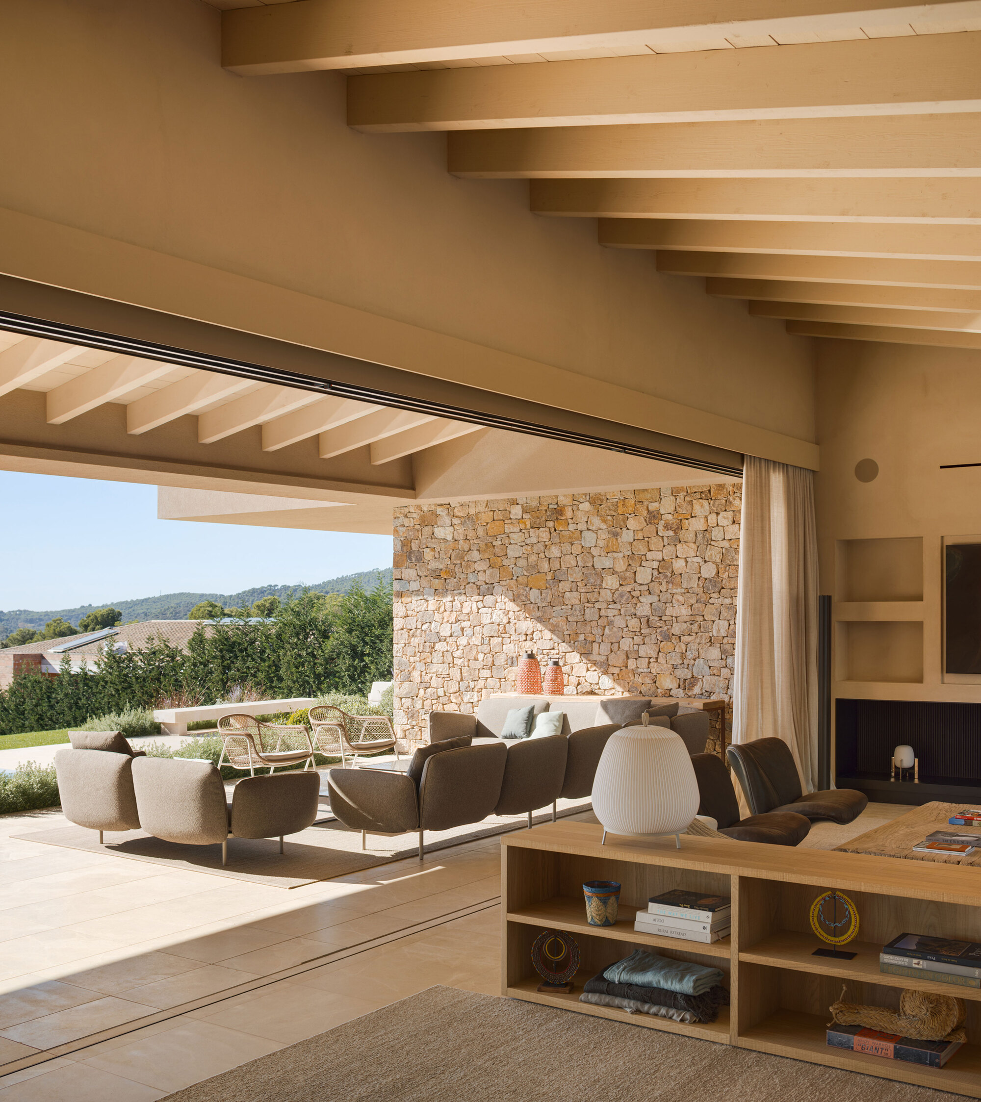 Open living room with timber beams, a rubble stone wall and a sliding glass wall facing wooded hills in afternoon sun