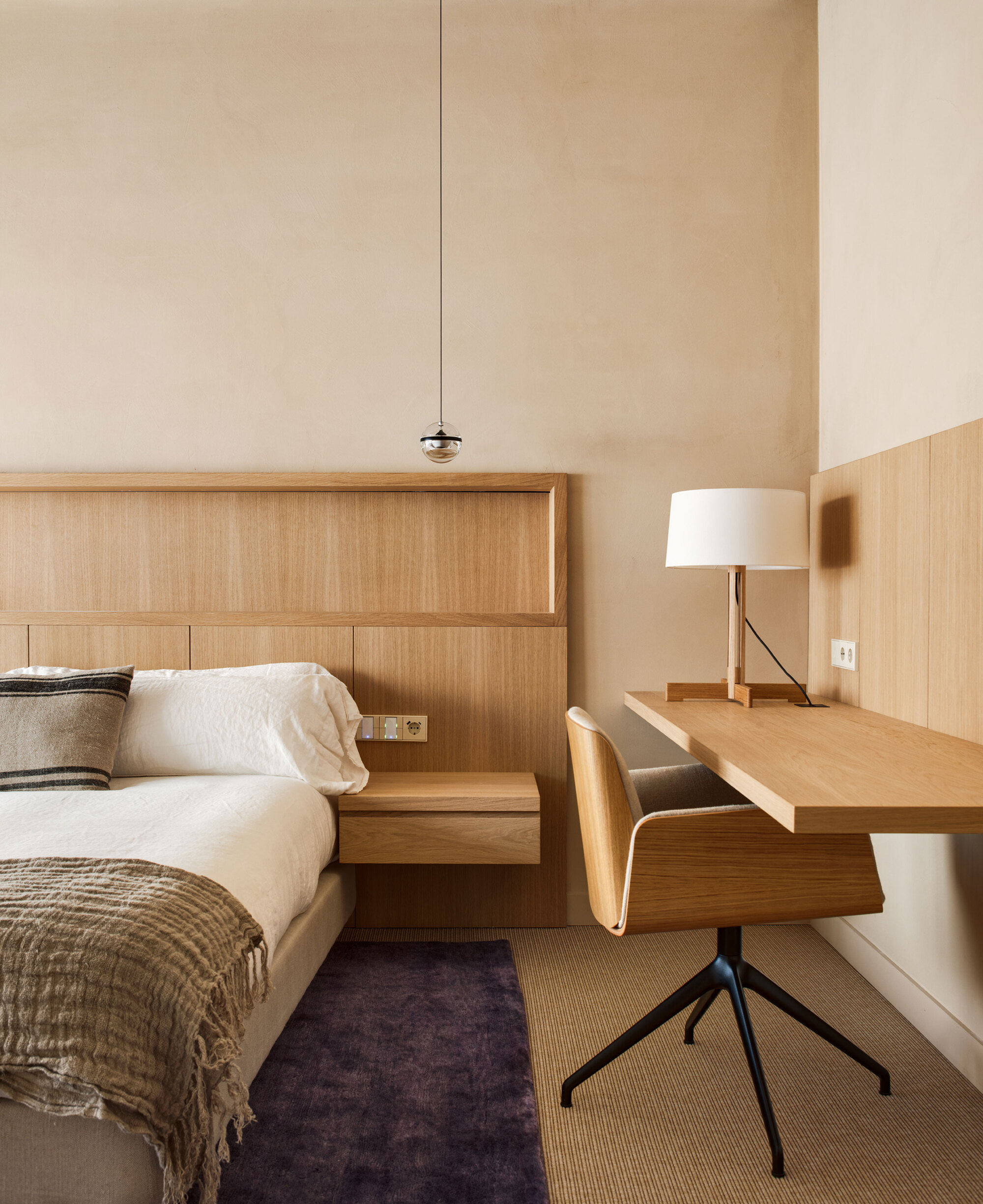 Bedroom corner with oak headboard, floating bedside shelf and wall-mounted desk beside a swivel chair and purple rug