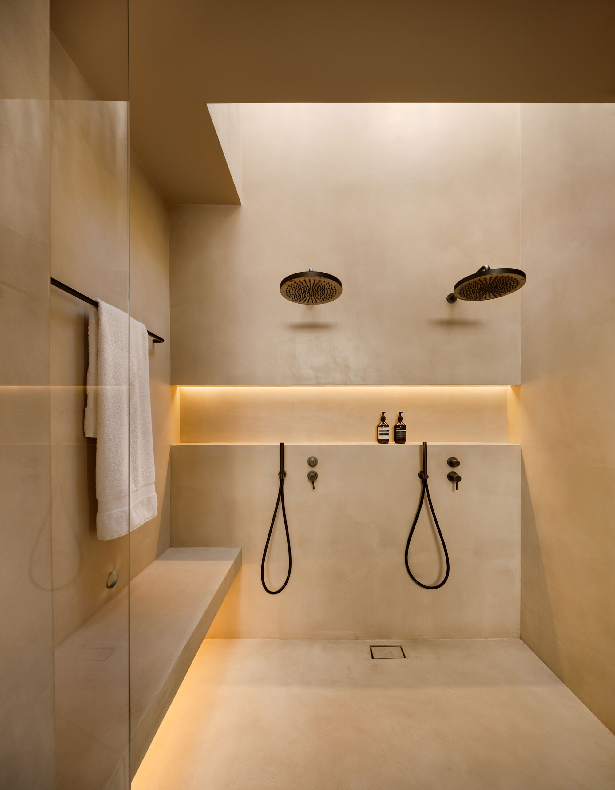 Walk-in shower with two round black rain heads, a lit niche and a built-in bench in plastered walls