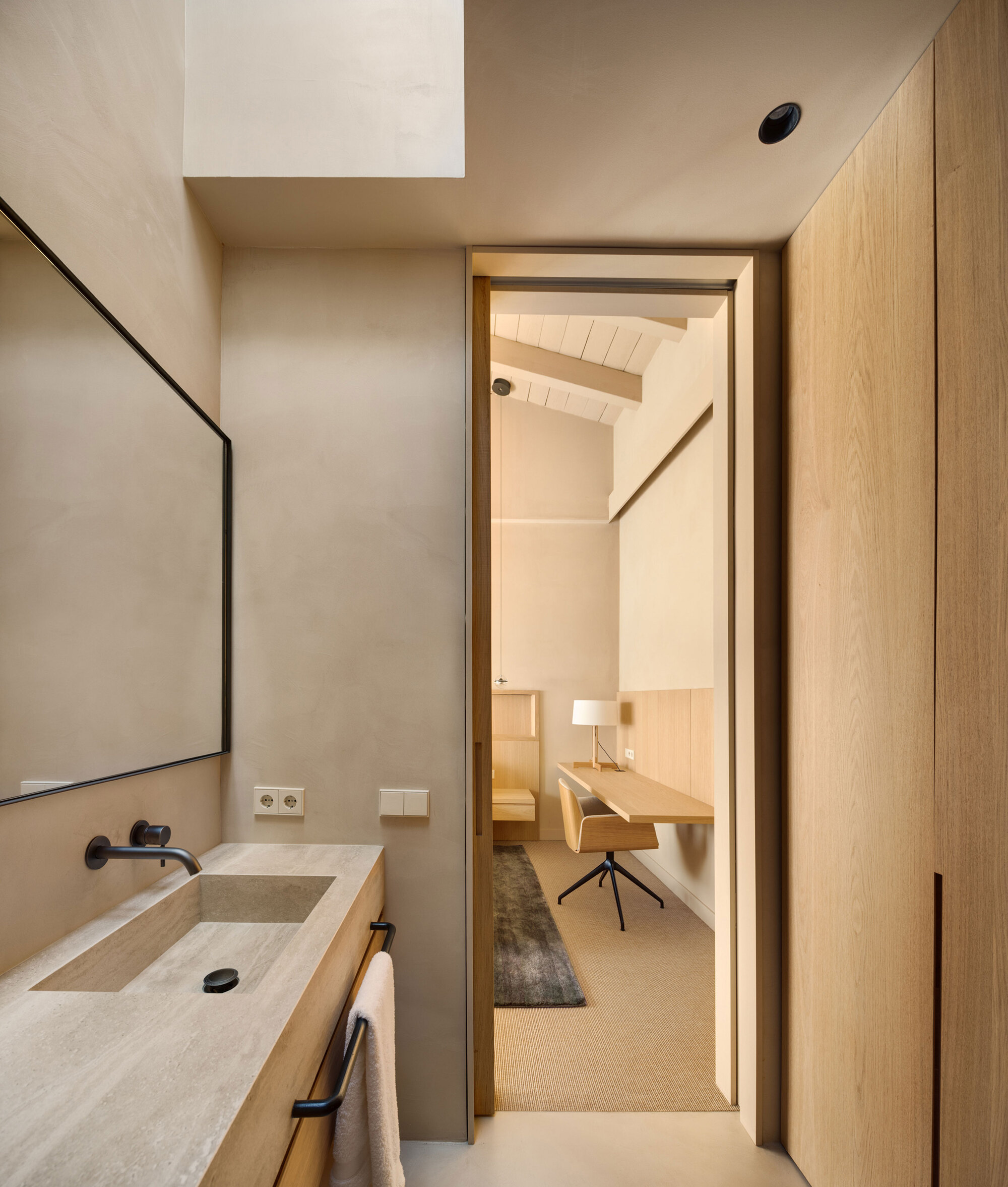Narrow bathroom with a stone basin and black framed mirror, a doorway opening to a bedroom desk under a pitched ceiling