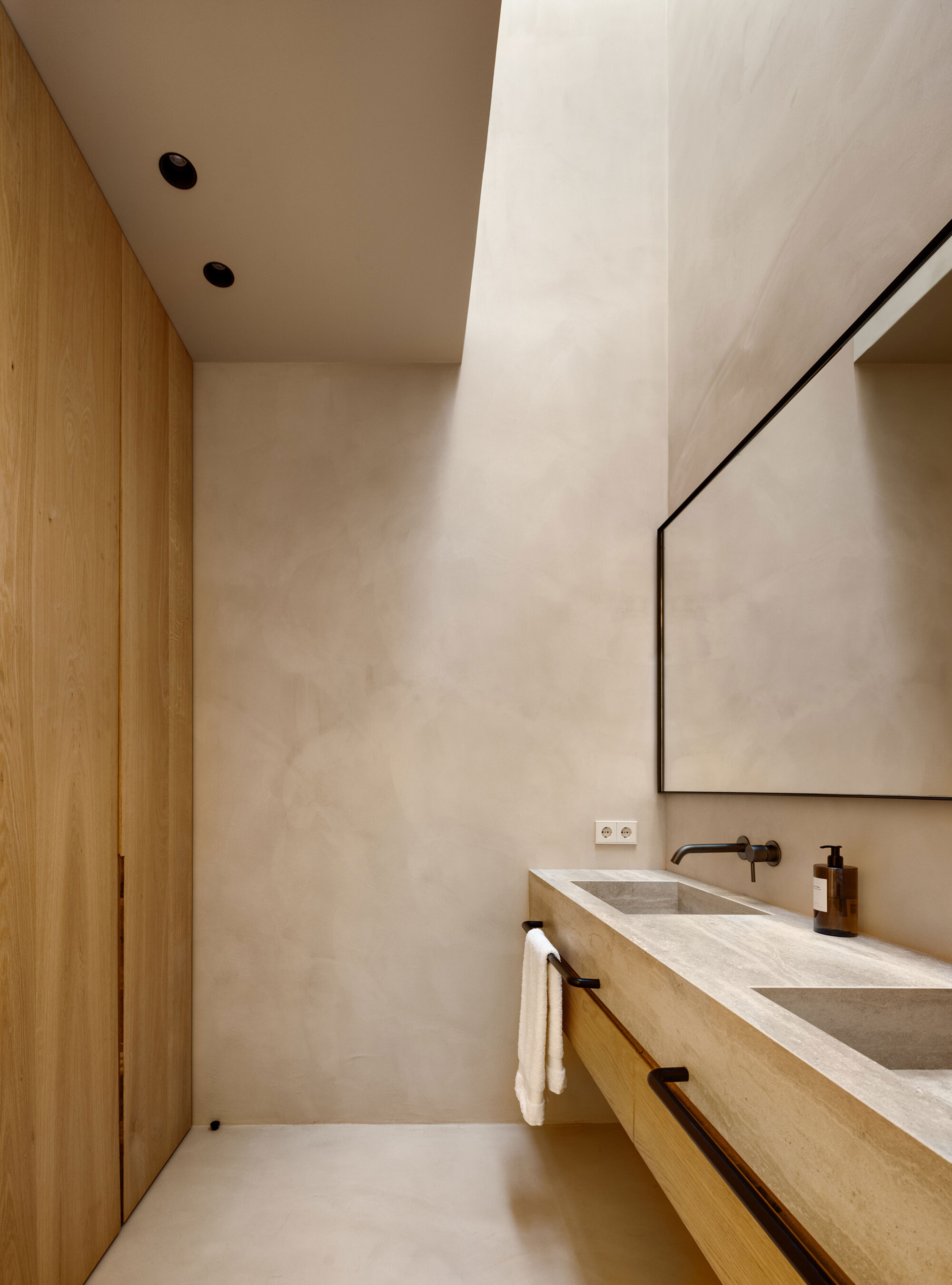 Bathroom with a floating travertine vanity, twin basins and oak cabinetry beside a tall skylight washing the plaster walls with daylight
