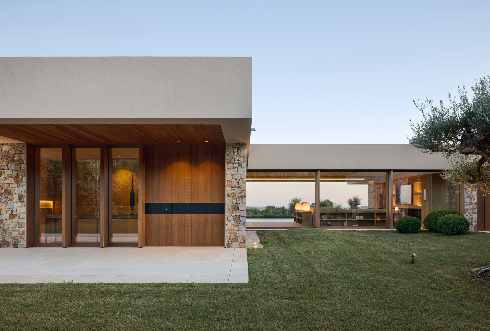 Entrance pavilion with stone piers, vertical timber door and flat roof beside an olive tree at dusk