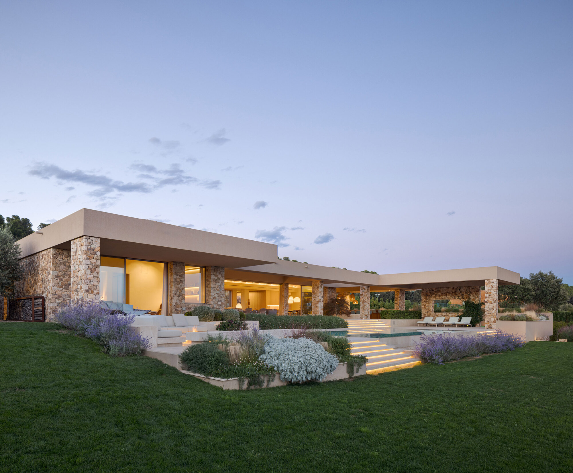 Low stone-pier house with a flat roof and lit pool steps behind lavender beds at twilight