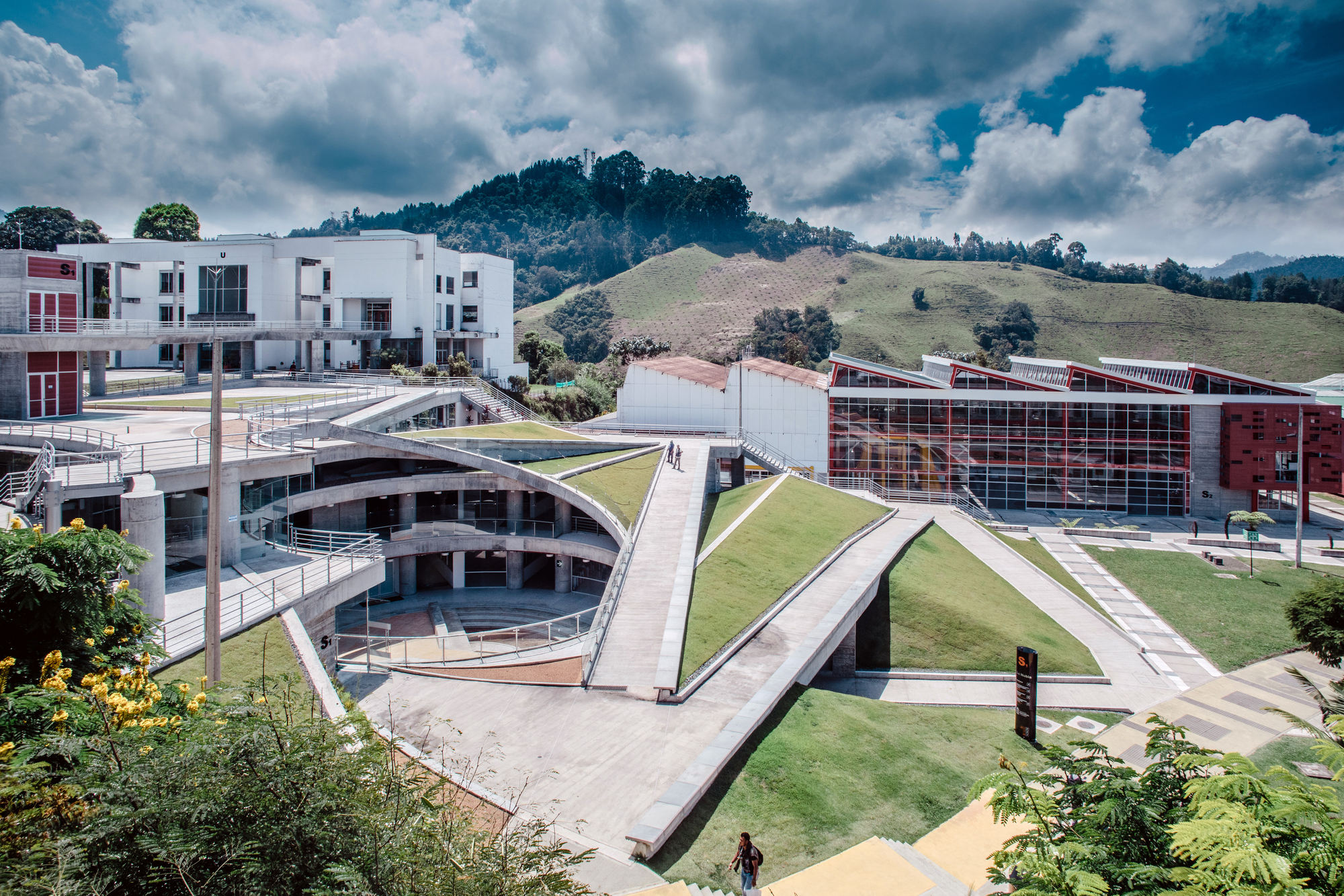 Grass-covered ramps and walkways descending around a circular amphitheater courtyard, with green hills and cumulus clouds behind