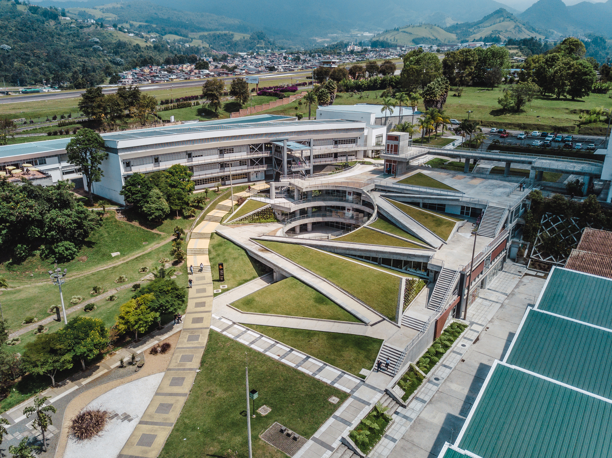 Aerial view of stepped grass terraces and ramps around a round courtyard, with teal metal roofs in bright sunlight