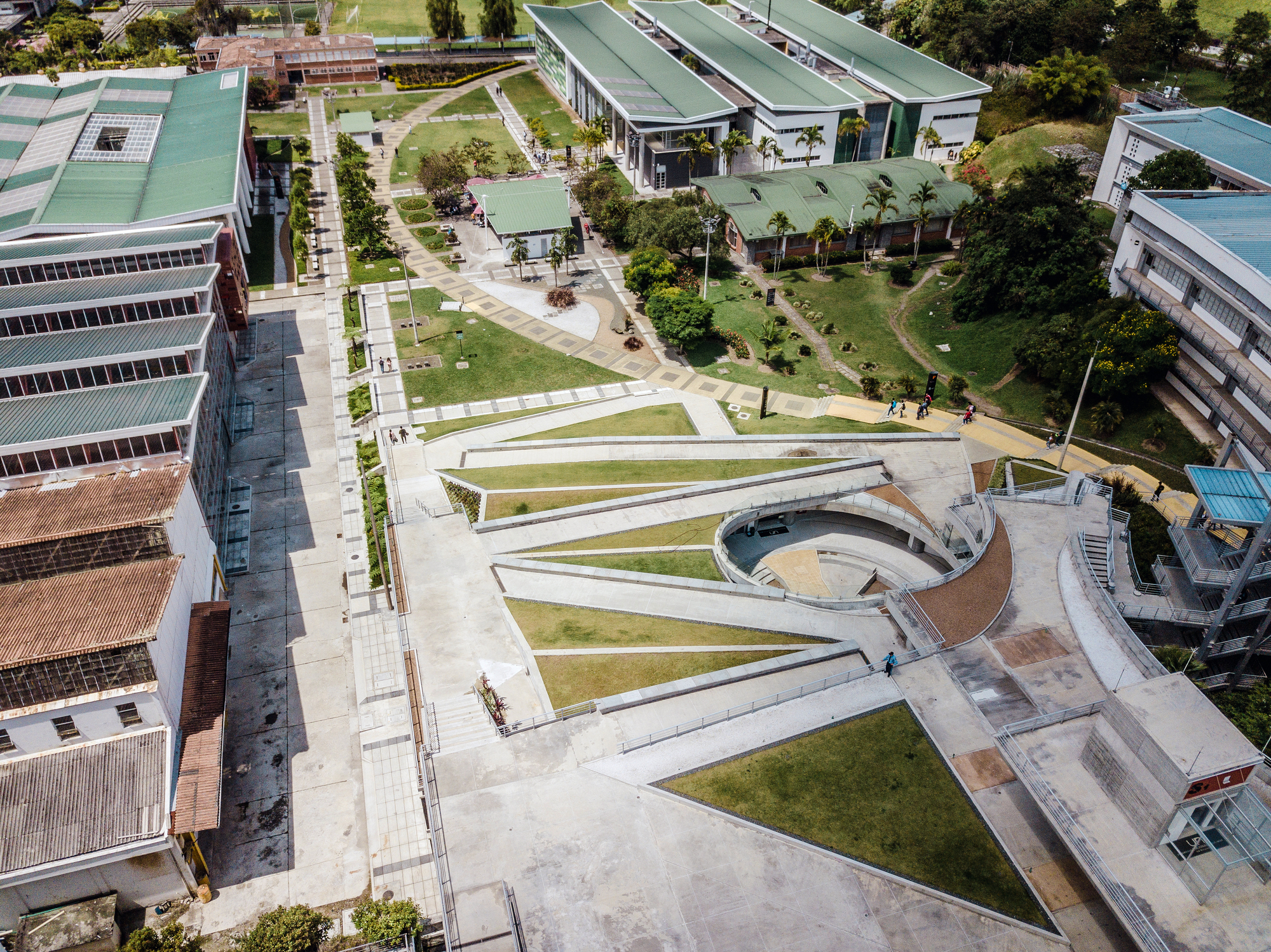 Top-down view of triangular lawns and concrete ramps beside a curved sunken court, with long roofed blocks and palms nearby