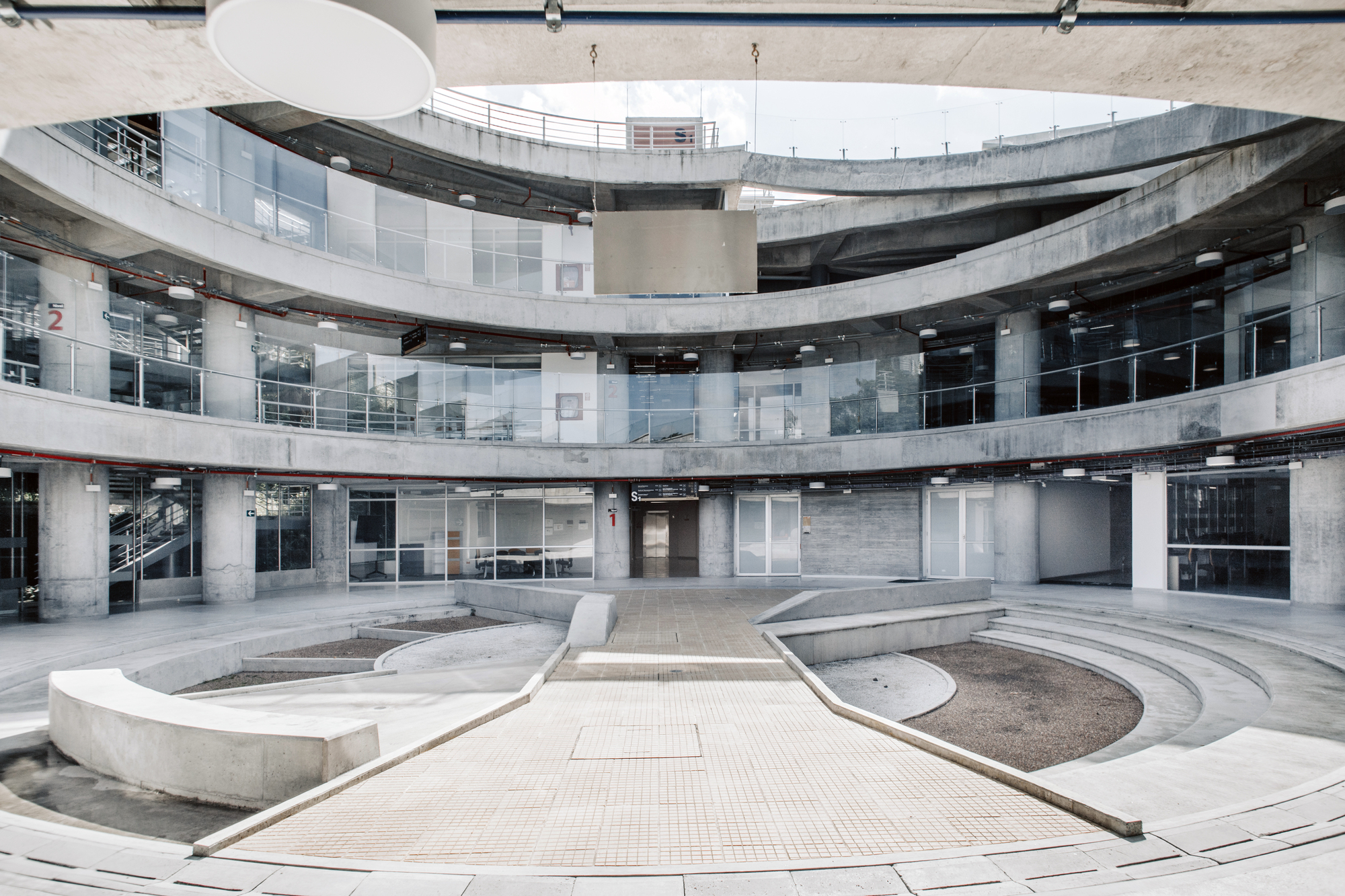 Curved concrete balconies ringing a tiled courtyard with planted beds and tiered seating, lit by soft daylight from above