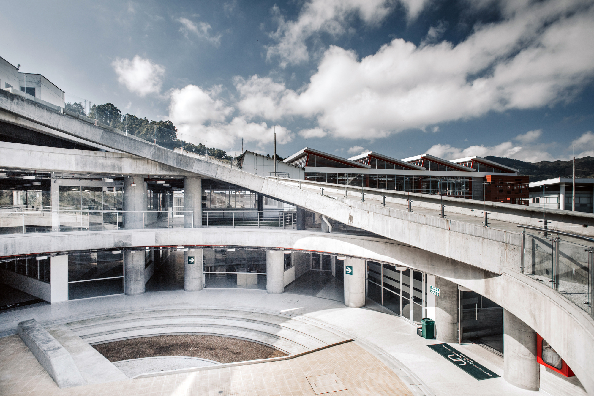 Sloping concrete ramp curving over round columns and a sunken seating court, under a sky of heavy white clouds