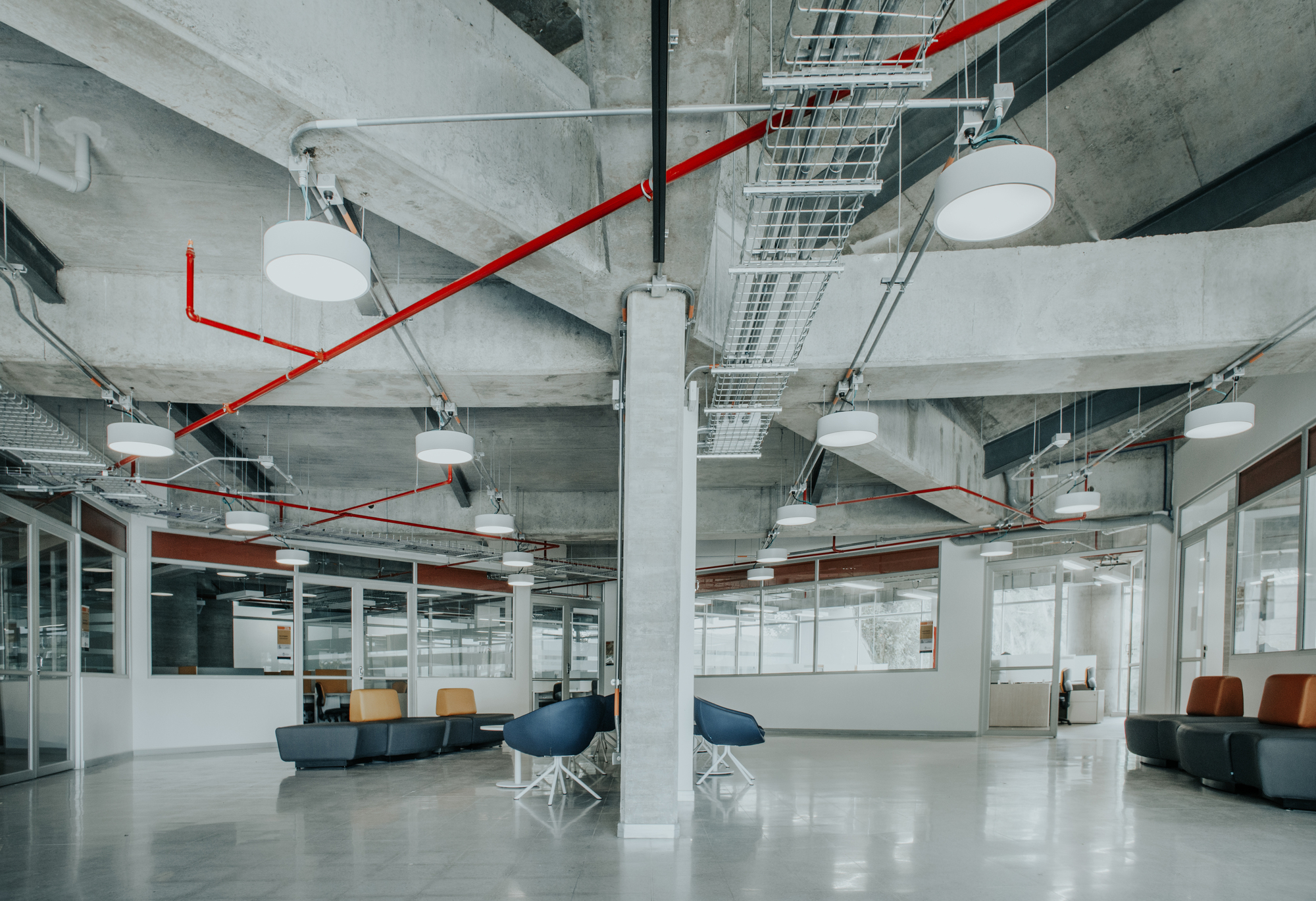 Exposed concrete beams and red sprinkler pipes above lounge seating and glazed offices, with round pendant lights hanging from the ceiling
