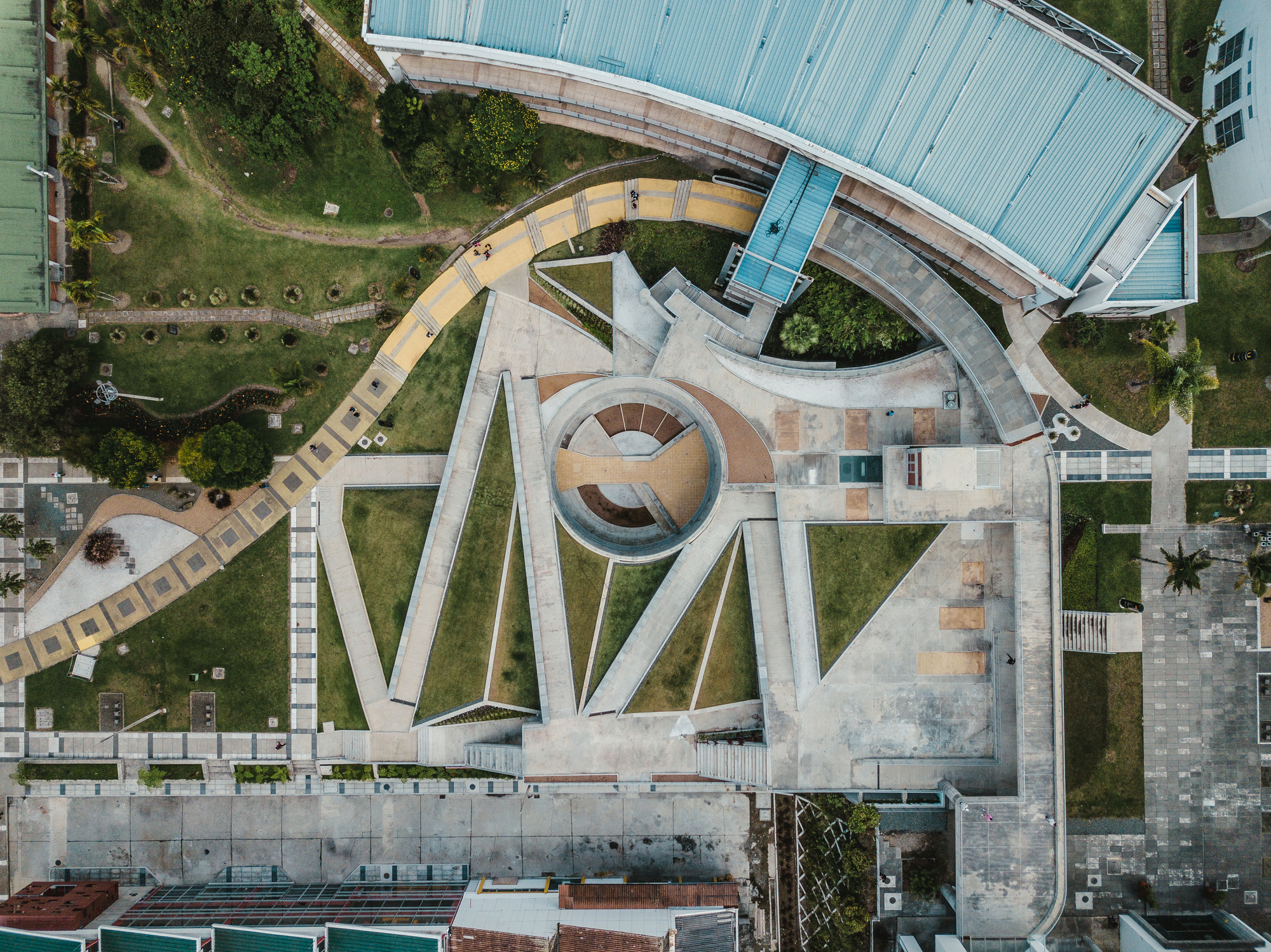 Drone view of a circular sunken court with radiating grass wedges and a curved yellow-tiled ramp across a green campus
