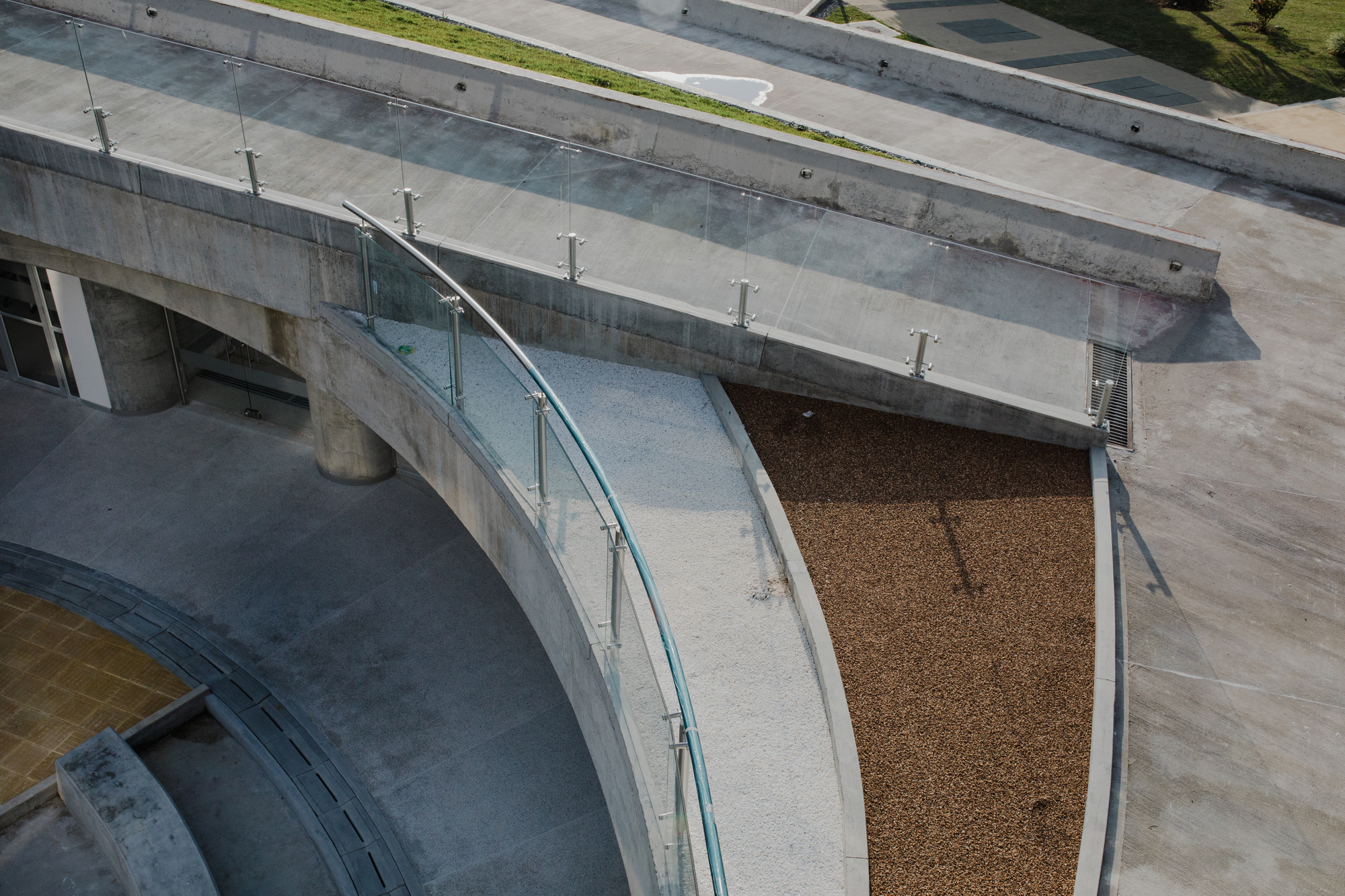 Curved concrete ramp with glass balustrade beside a bed of brown gravel, seen from above in flat daylight