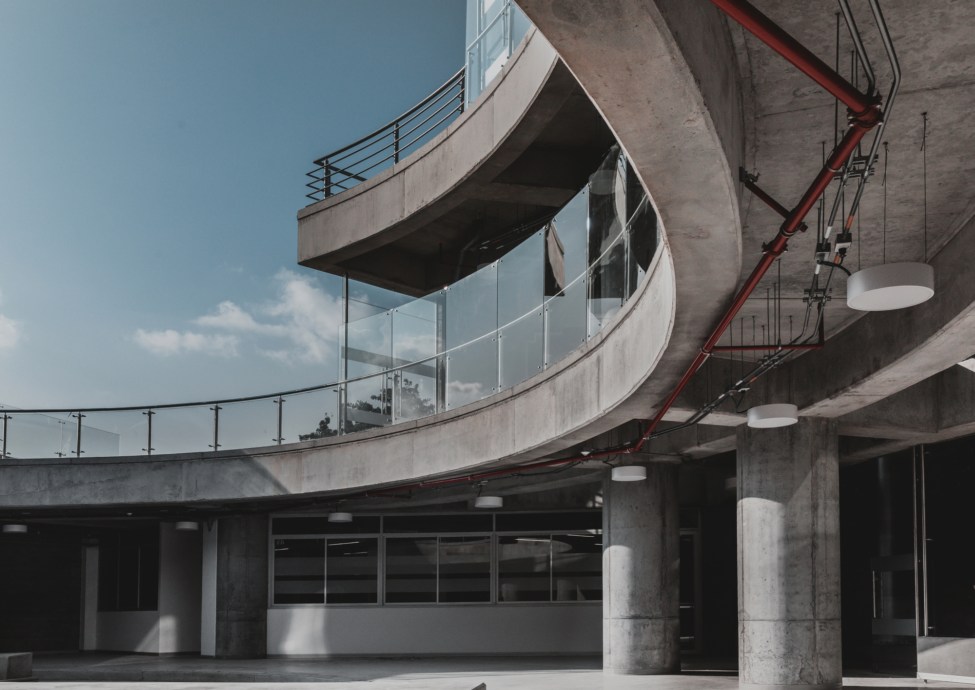 Curving concrete balconies with glass railings and a red pipe running along the soffit beneath a blue sky with thin clouds