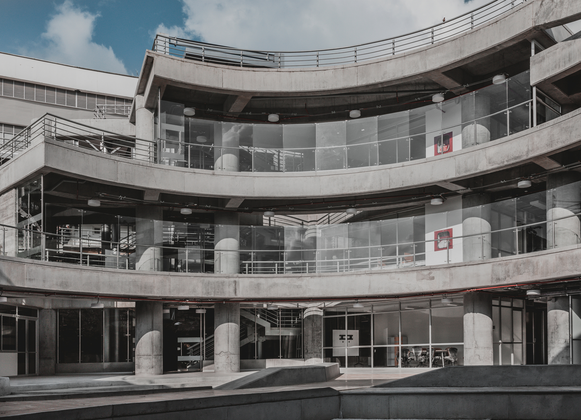 Stacked curved concrete floors with glass guardrails around a courtyard, round columns at ground level under a cloudy sky