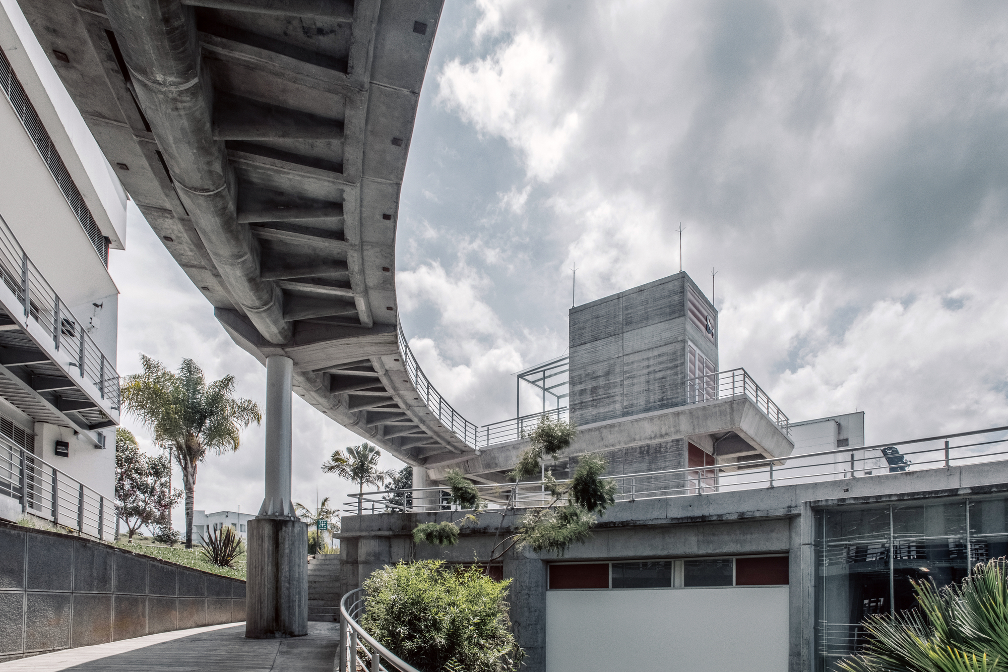 Underside of a sweeping concrete ramp on a single round column, with palms and a concrete tower under heavy clouds