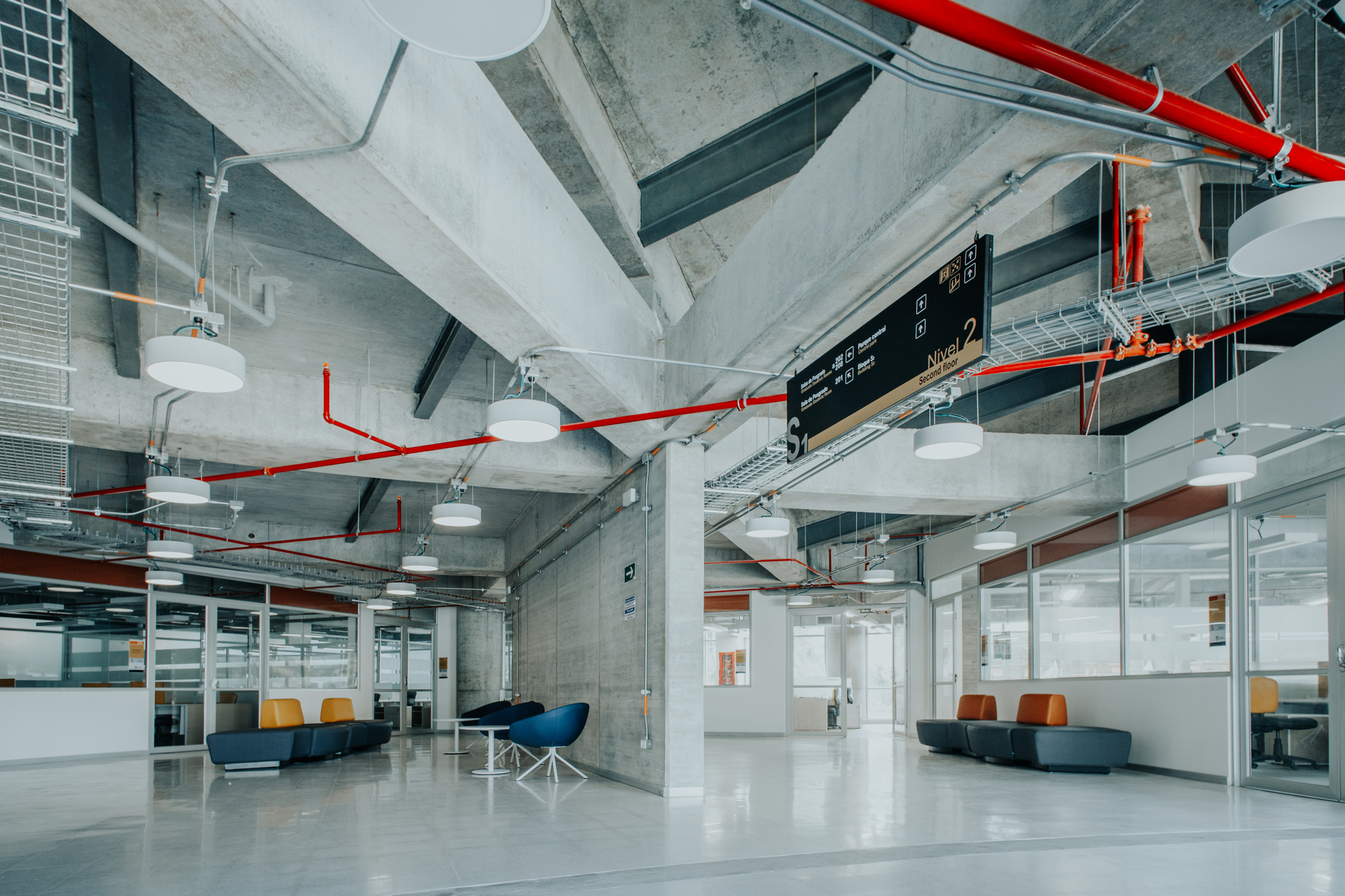 Concrete-ceilinged lounge with red sprinkler pipes, round pendant lights and blue and orange seating beside glazed partitions