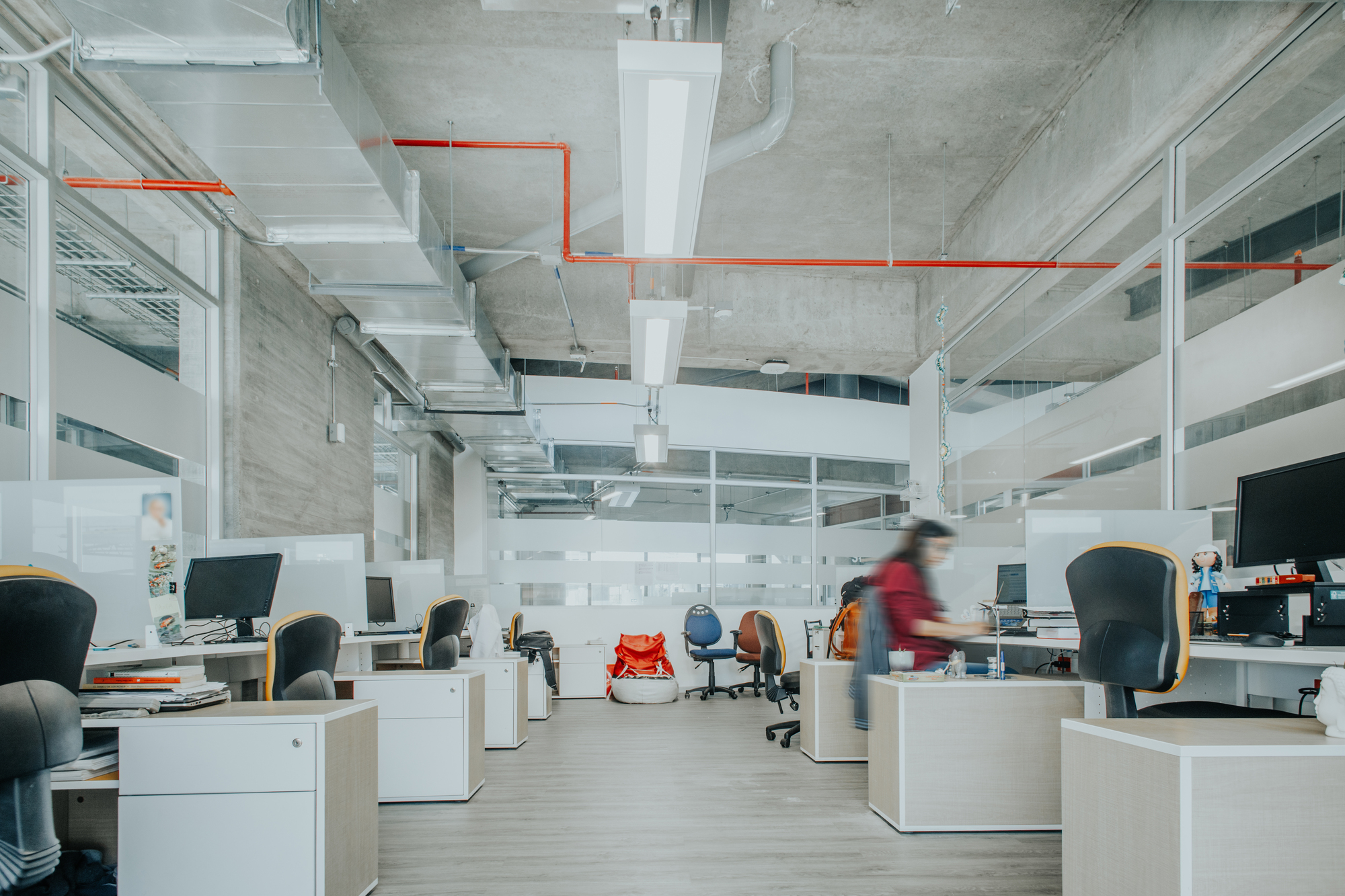 Open office with rows of white desks, exposed concrete ceiling and a blurred worker in red at her desk