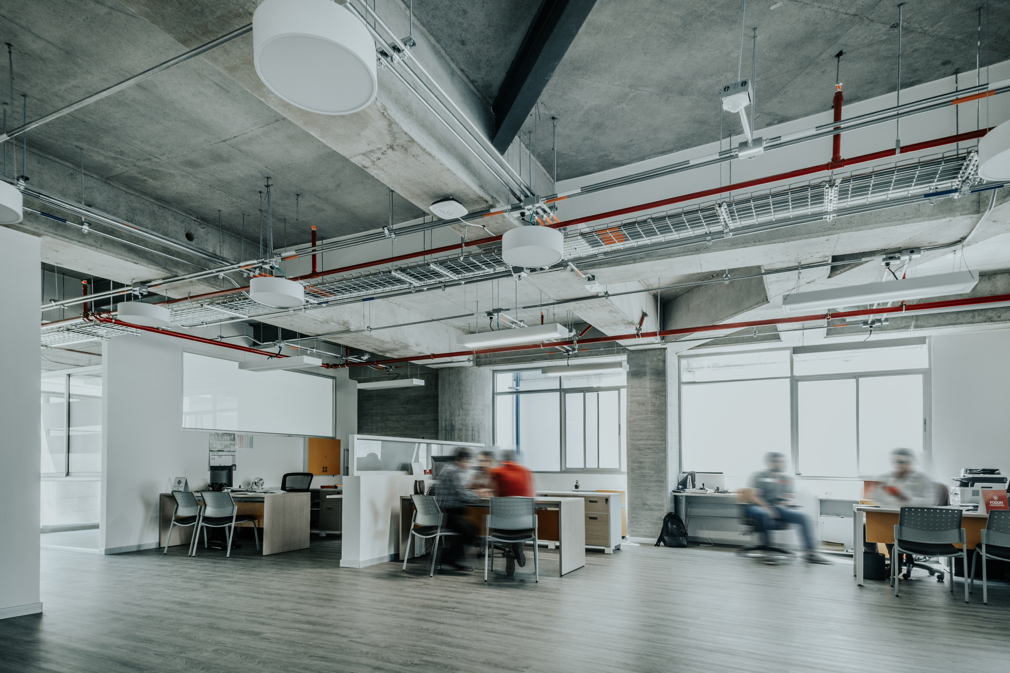 Open workspace under a concrete beam ceiling with red pipes, cable trays and blurred colleagues meeting around a table