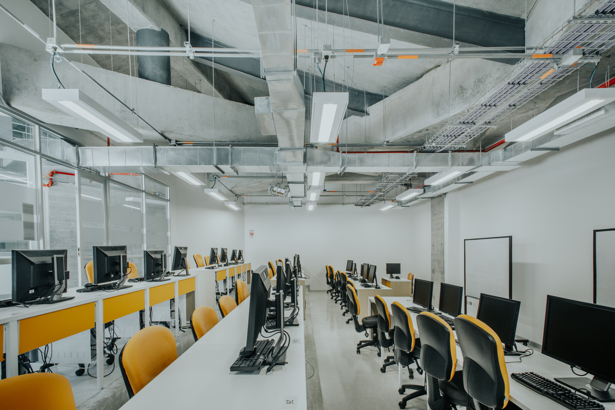 Computer lab with long white desks, yellow chairs and monitors beneath exposed ducts and linear light fixtures