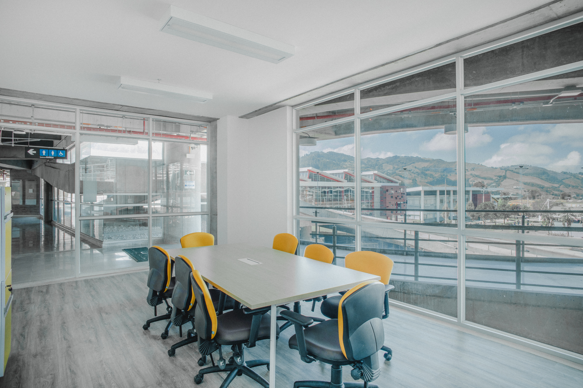 Meeting room with a long table and yellow chairs facing a glass wall that looks onto green mountains and cloud