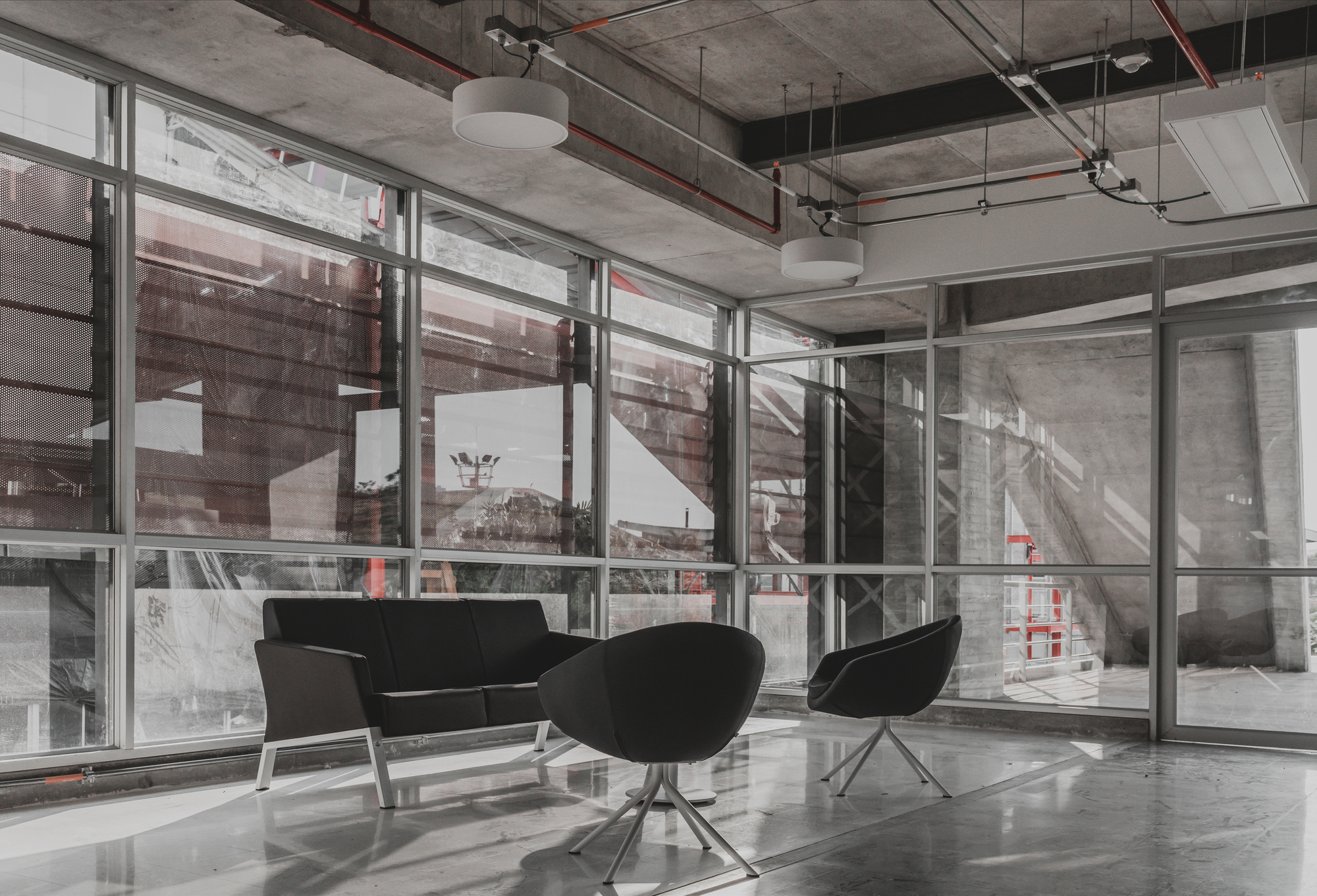 Lounge corner with a black sofa and two bucket chairs before a glazed wall, exposed concrete ceiling and red conduits above