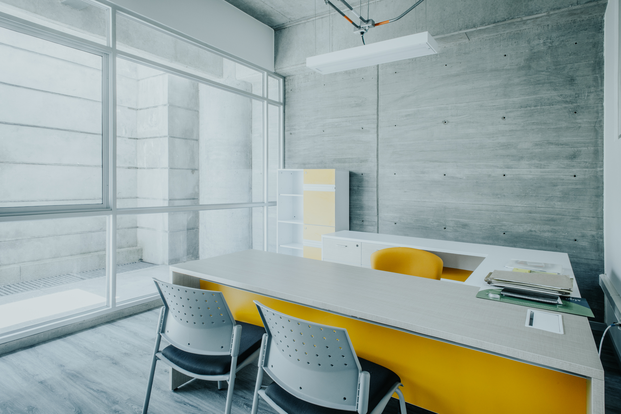 Private office with a yellow-fronted desk and board-marked concrete wall beside a full-height window facing a stepped concrete light well