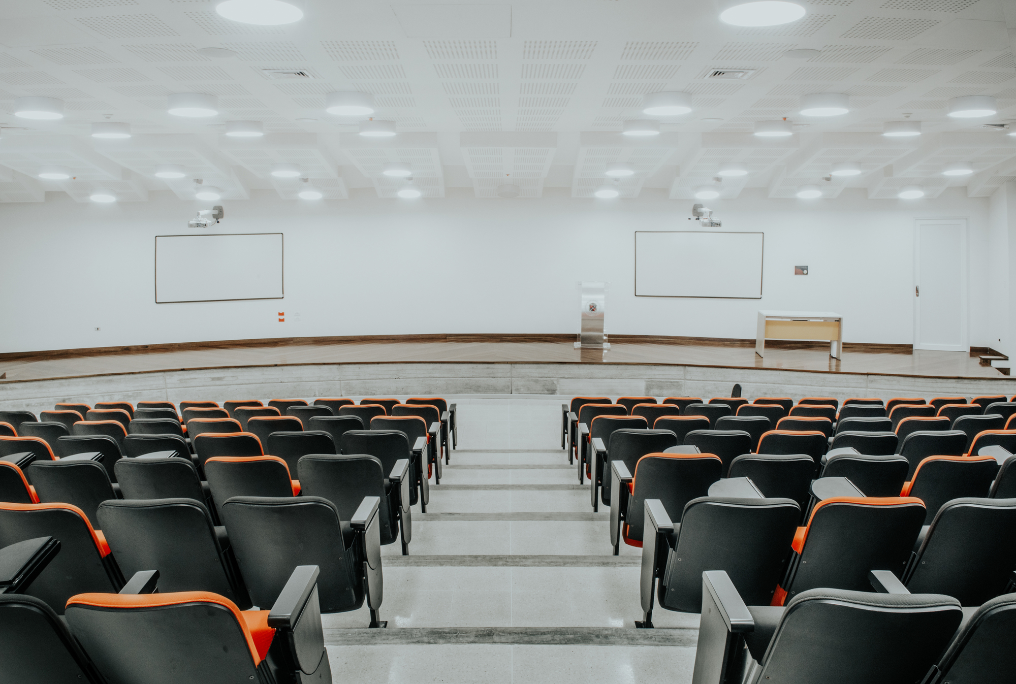 Tiered lecture hall with black and orange seats facing two whiteboards under a ceiling of recessed round lights