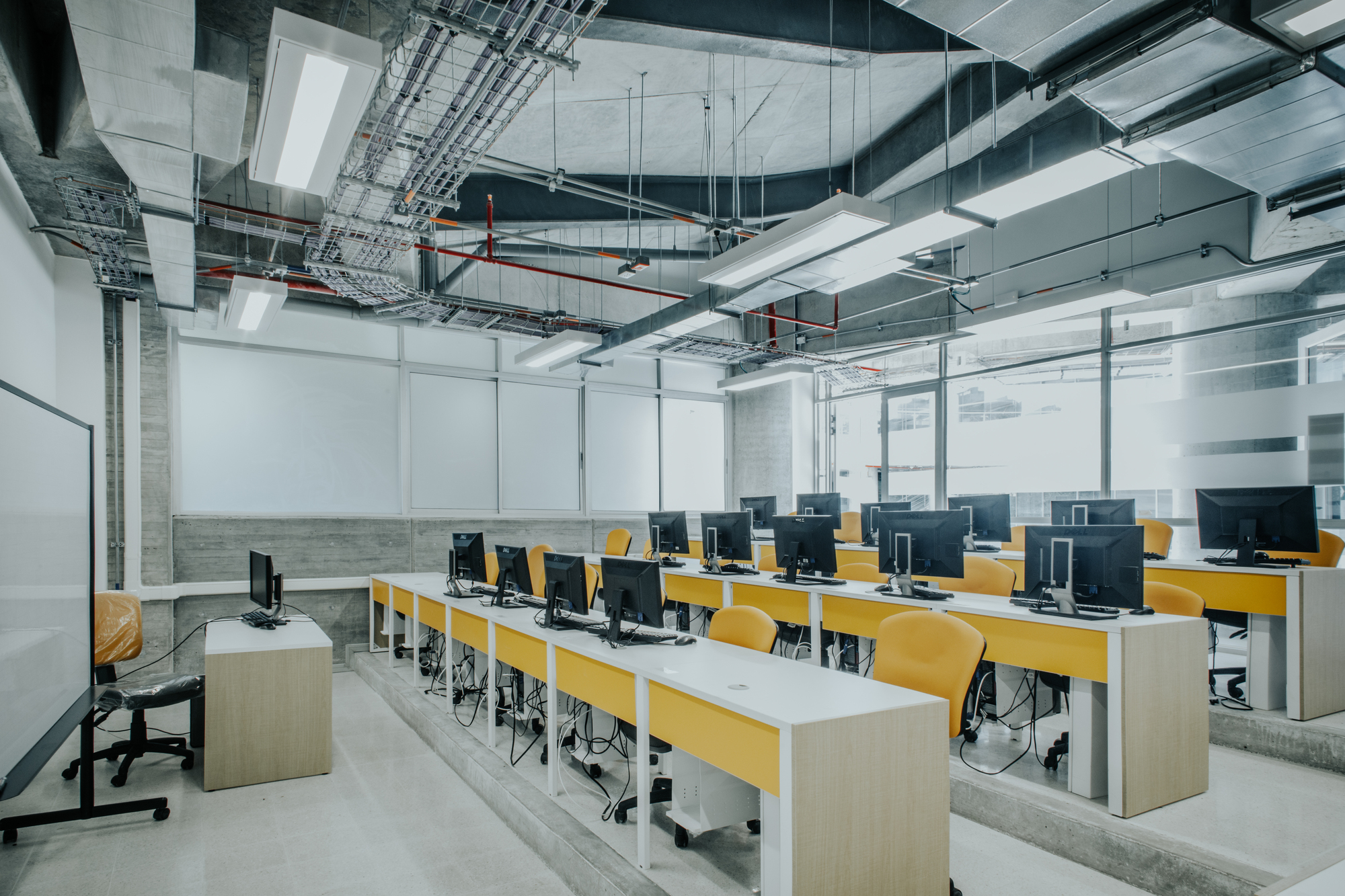 Computer classroom with rows of yellow-fronted desks on a stepped floor beneath exposed ducts, red pipes and fluorescent fittings