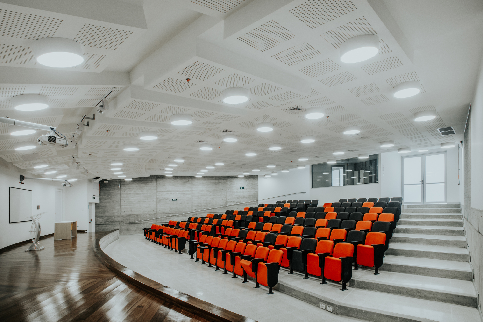 Tiered lecture hall with orange and black seats, concrete side wall and perforated ceiling panels lit by round lamps over a timber floor