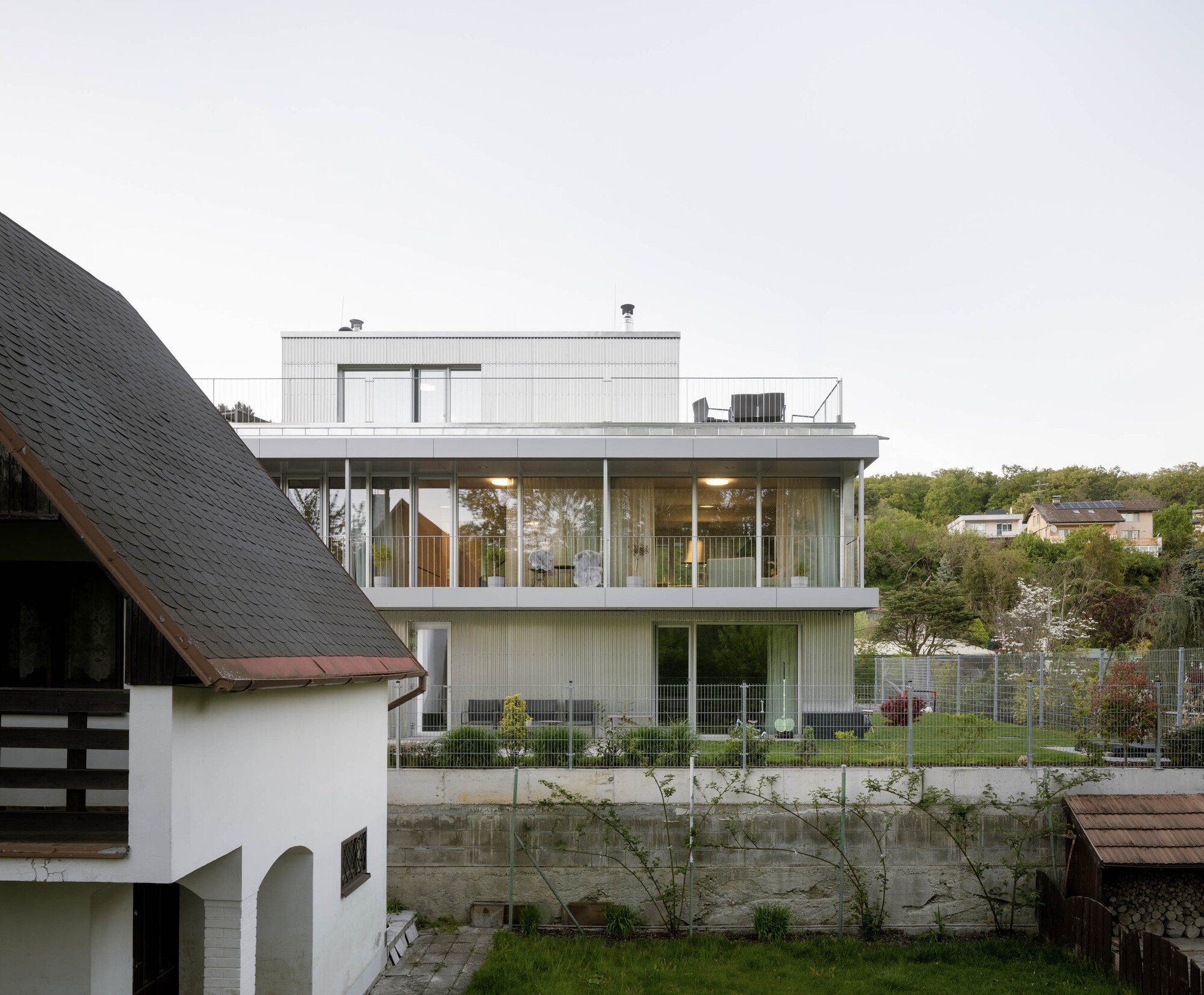 Stepped glazed volume with a rooftop terrace behind a retaining wall and a neighbouring gabled house at dusk