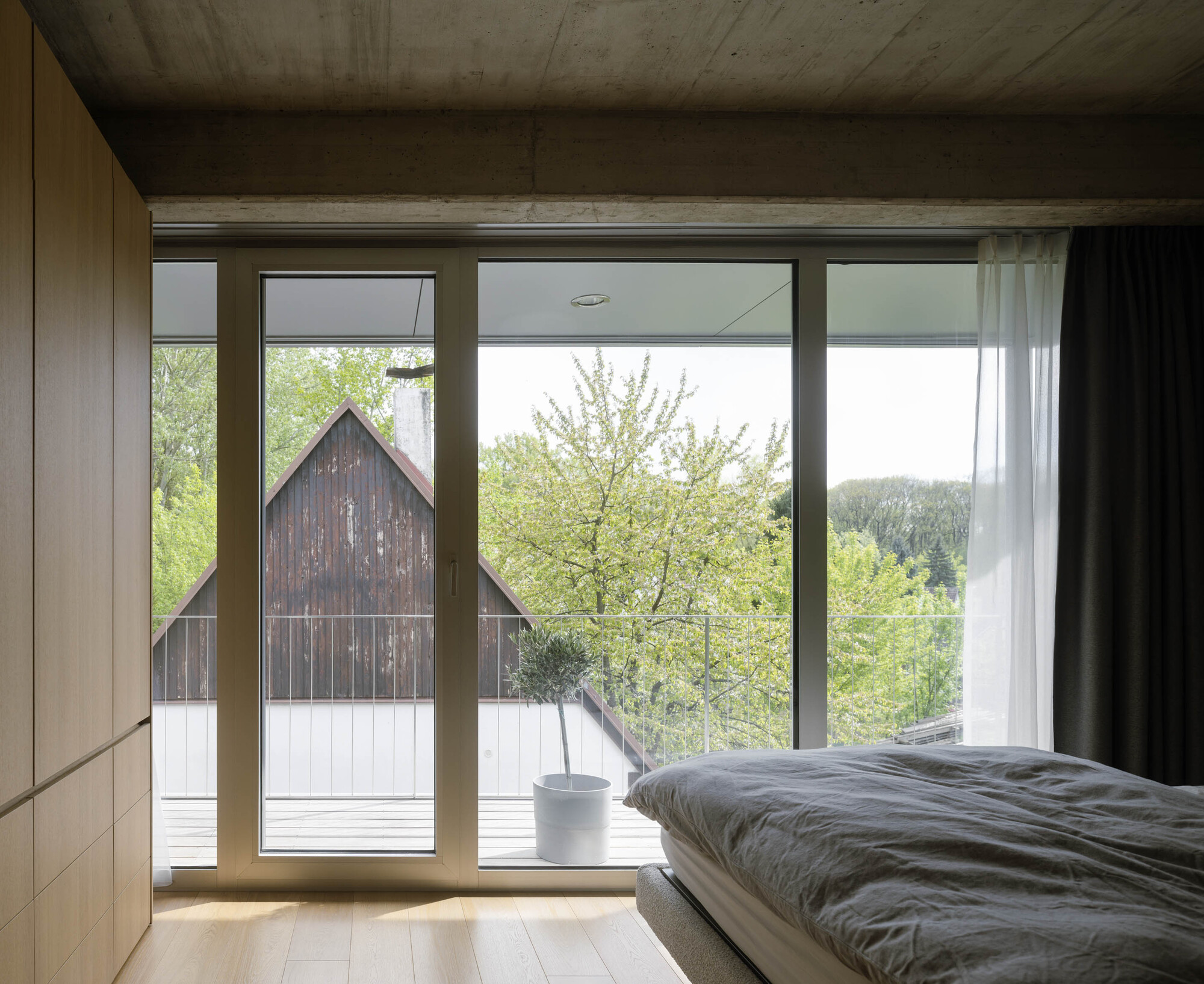 Bedroom with concrete ceiling, oak wardrobe and floor-to-ceiling glazing opening onto a terrace facing a timber gabled barn