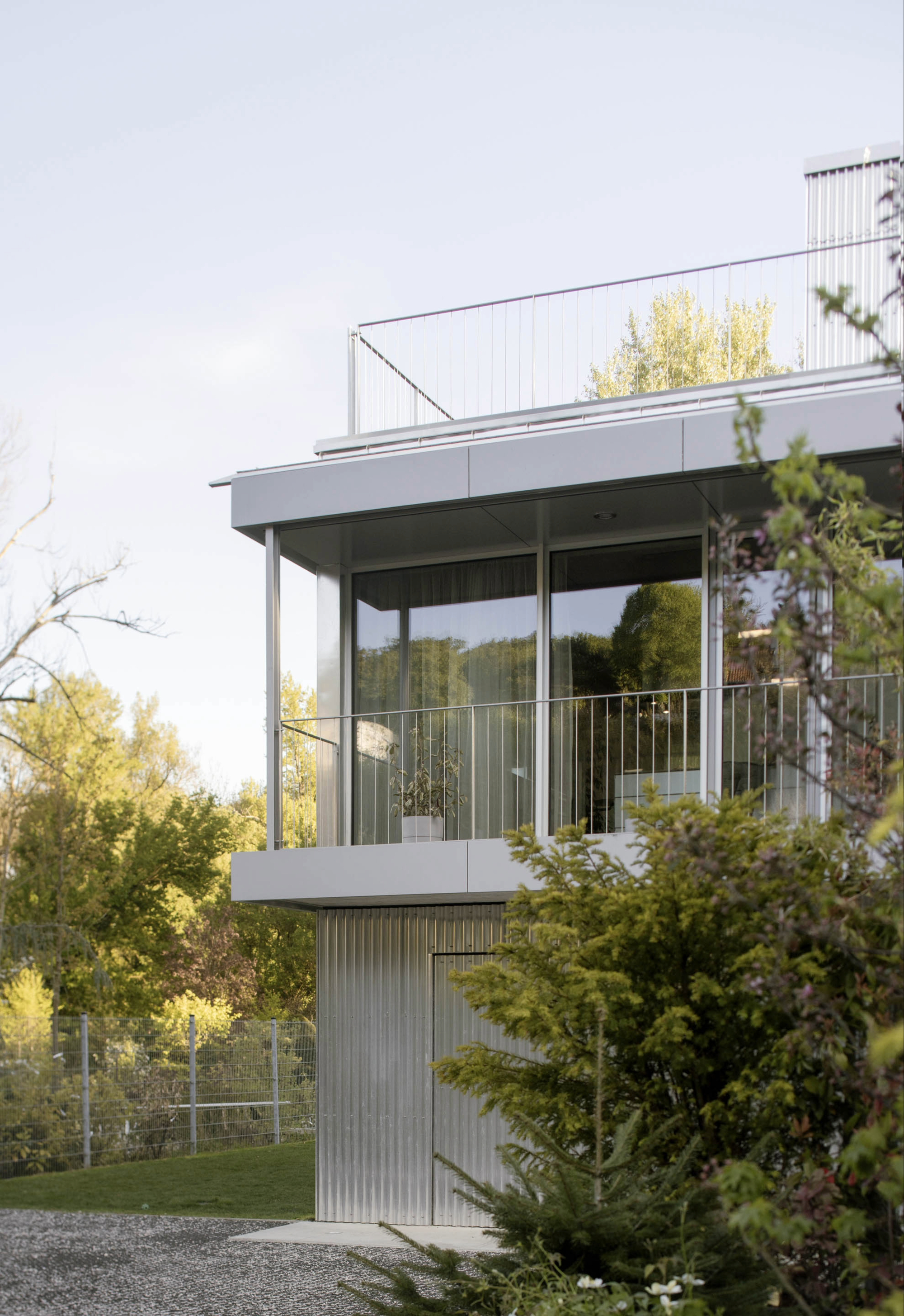 Corner of the upper floor with glazed balcony, metal railing and corrugated metal cladding below, framed by evergreen branches in soft evening light