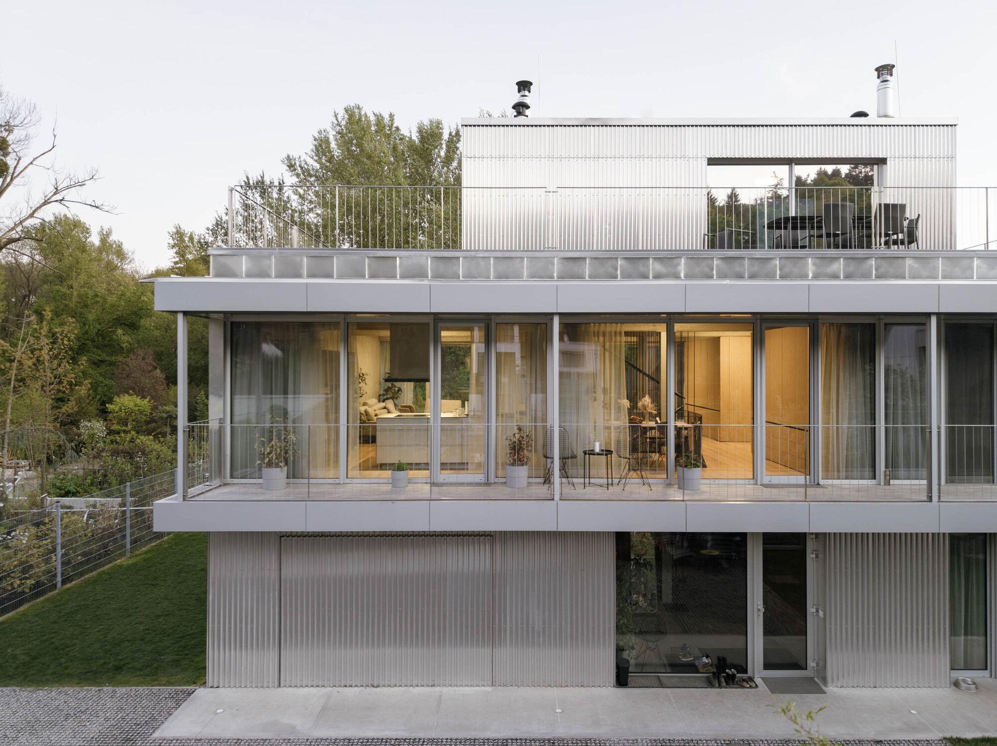 Front facade with a glazed balcony floor above corrugated metal garage cladding and a rooftop terrace, beside a grassy slope at dusk