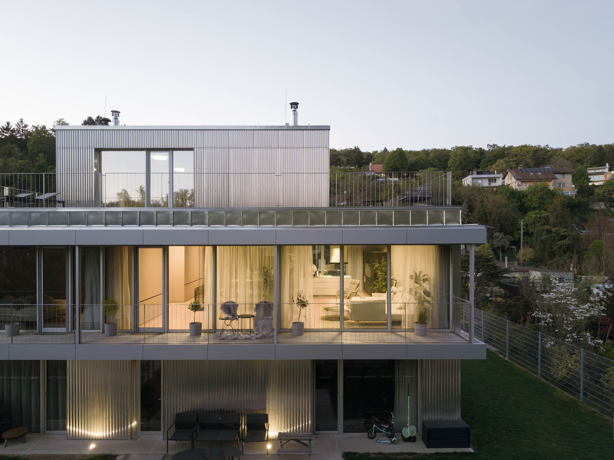 Side view of the stacked balconies and glazed living floor, with lit corrugated walls at ground level and wooded hillside houses beyond