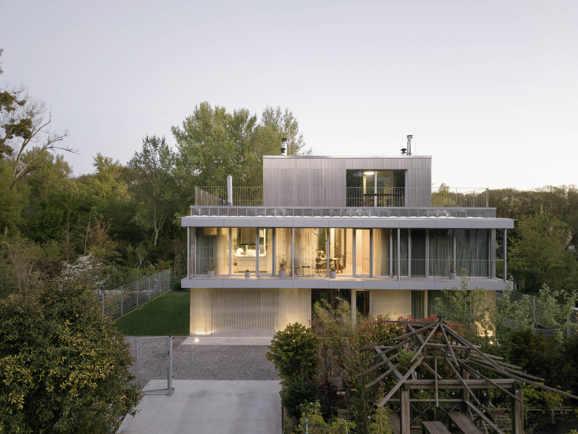 Frontal view of the three-level house with rooftop volume, set among trees and a timber pergola in the foreground at dusk