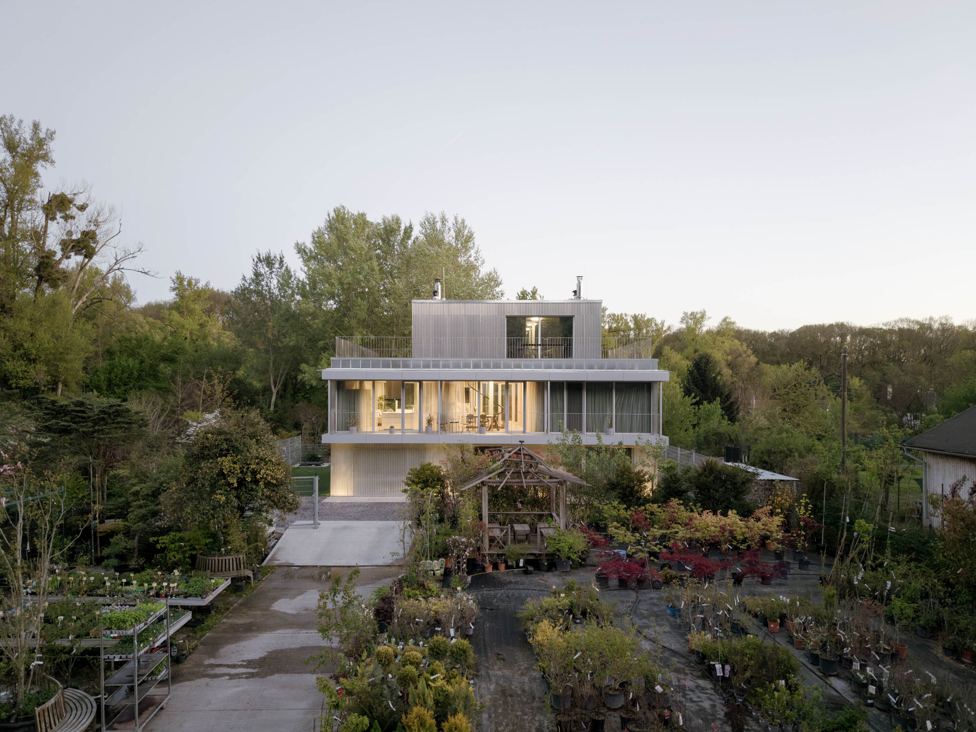 Distant view of the house rising above a plant nursery with potted shrubs, a wooden gazebo and a wet gravel path
