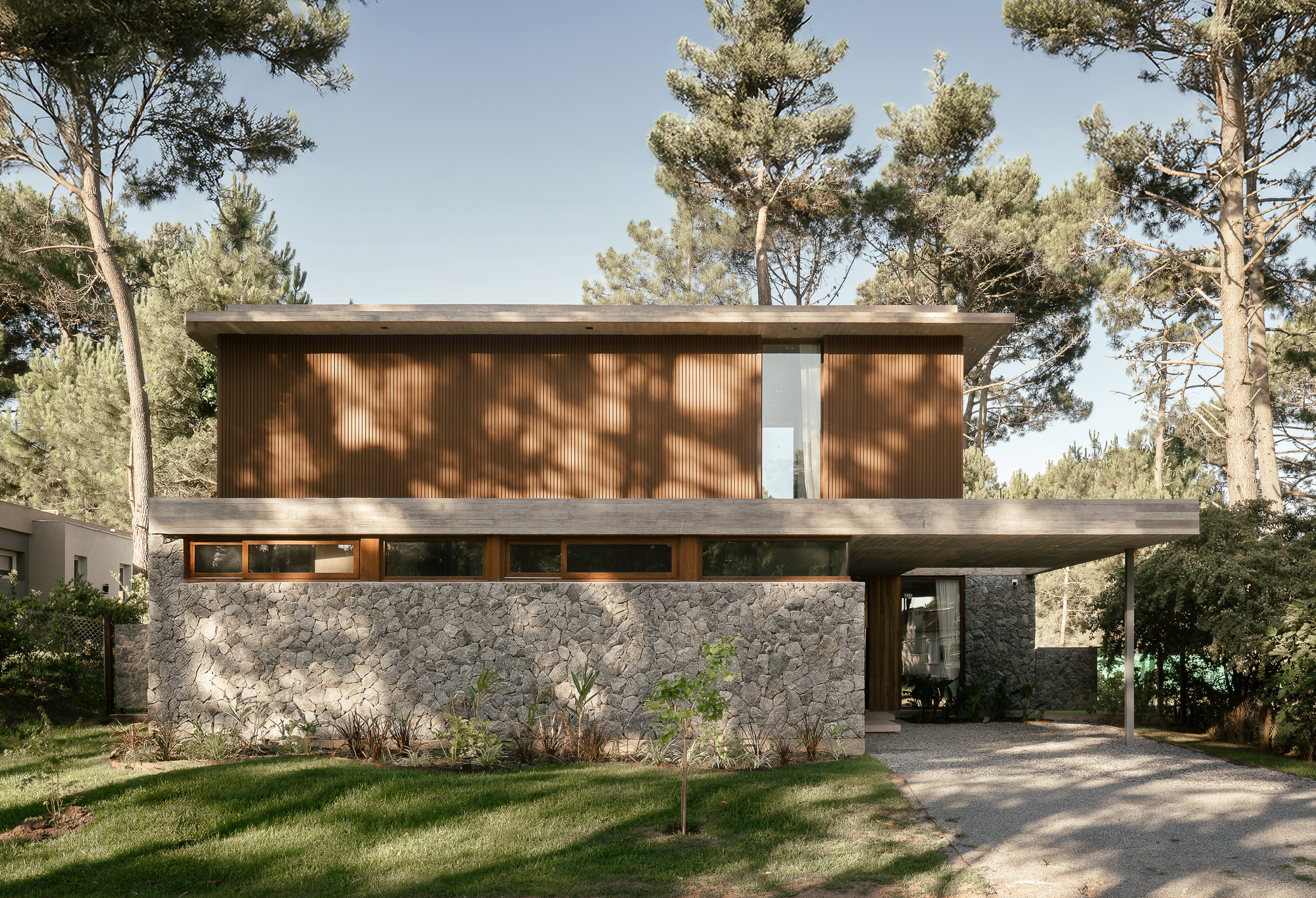 Street facade with a timber-clad upper volume above a rubble stone base and concrete canopy, dappled by pine shadows