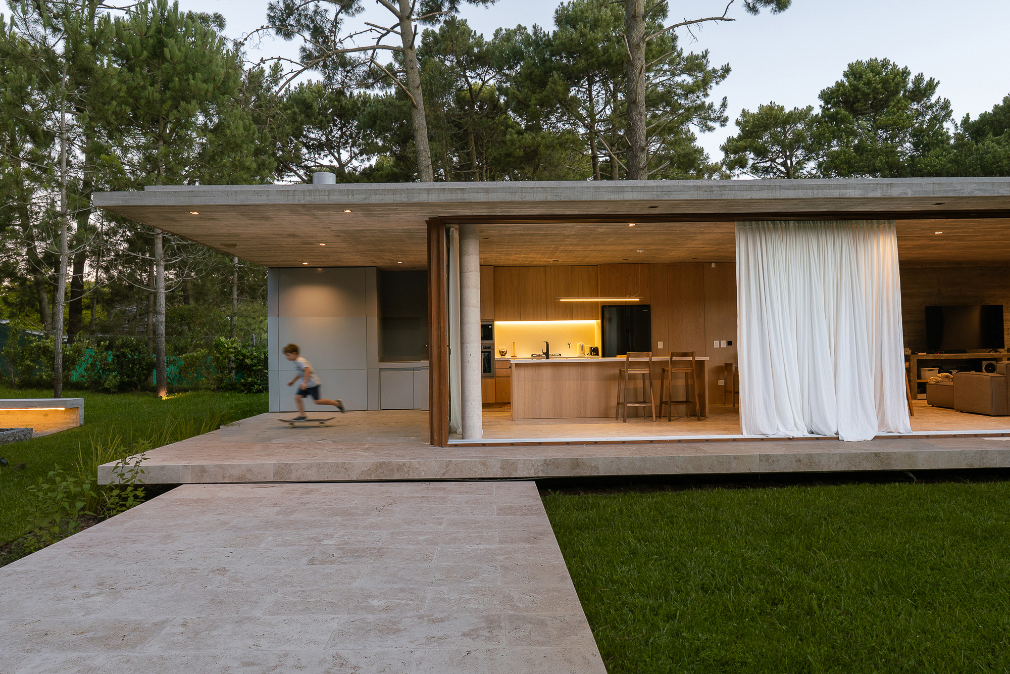 Open kitchen pavilion with white curtain and travertine path at dusk, a child skateboarding across the terrace
