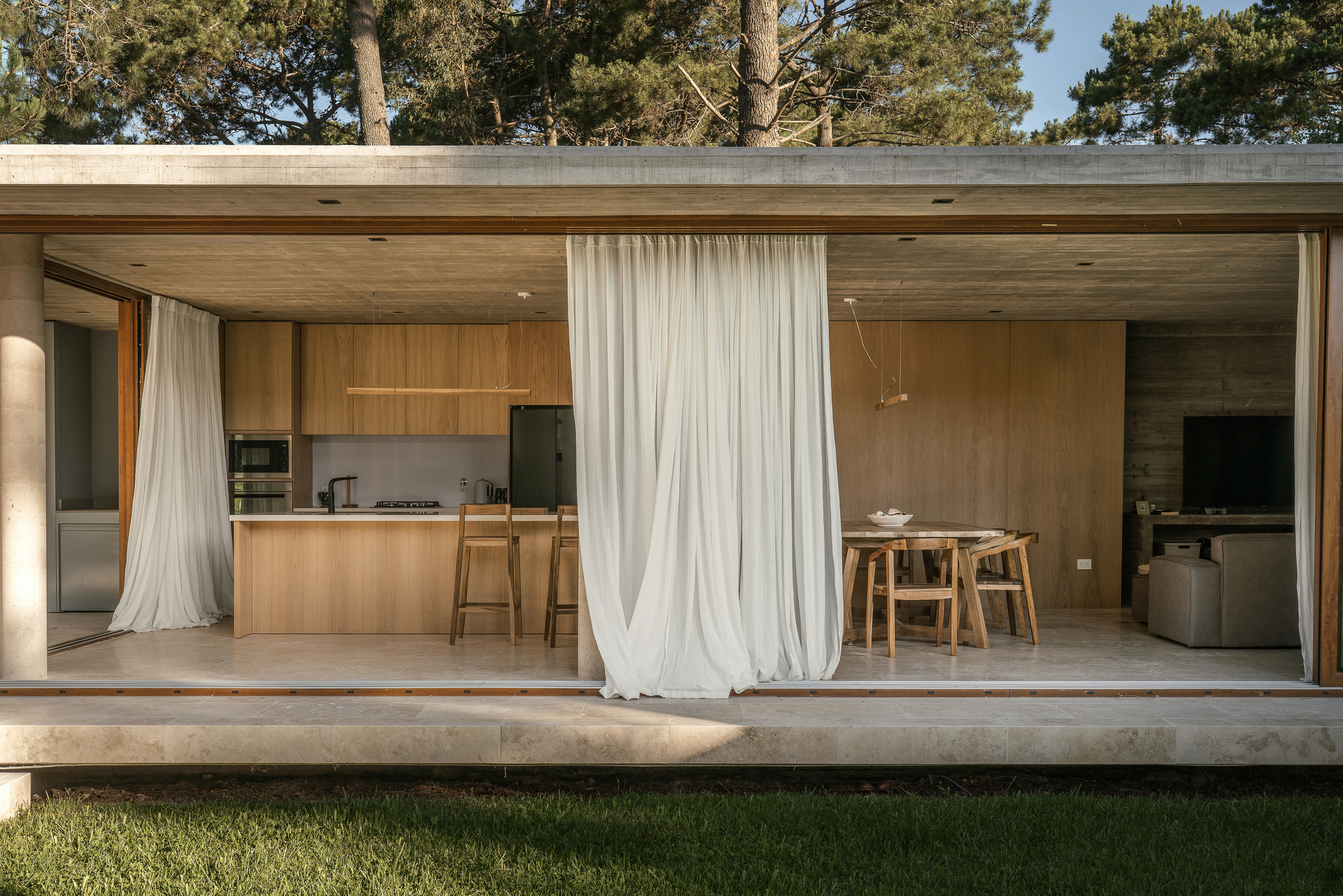 Kitchen and dining area framed by a concrete slab roof and sheer curtains, seen across the lawn in low sun