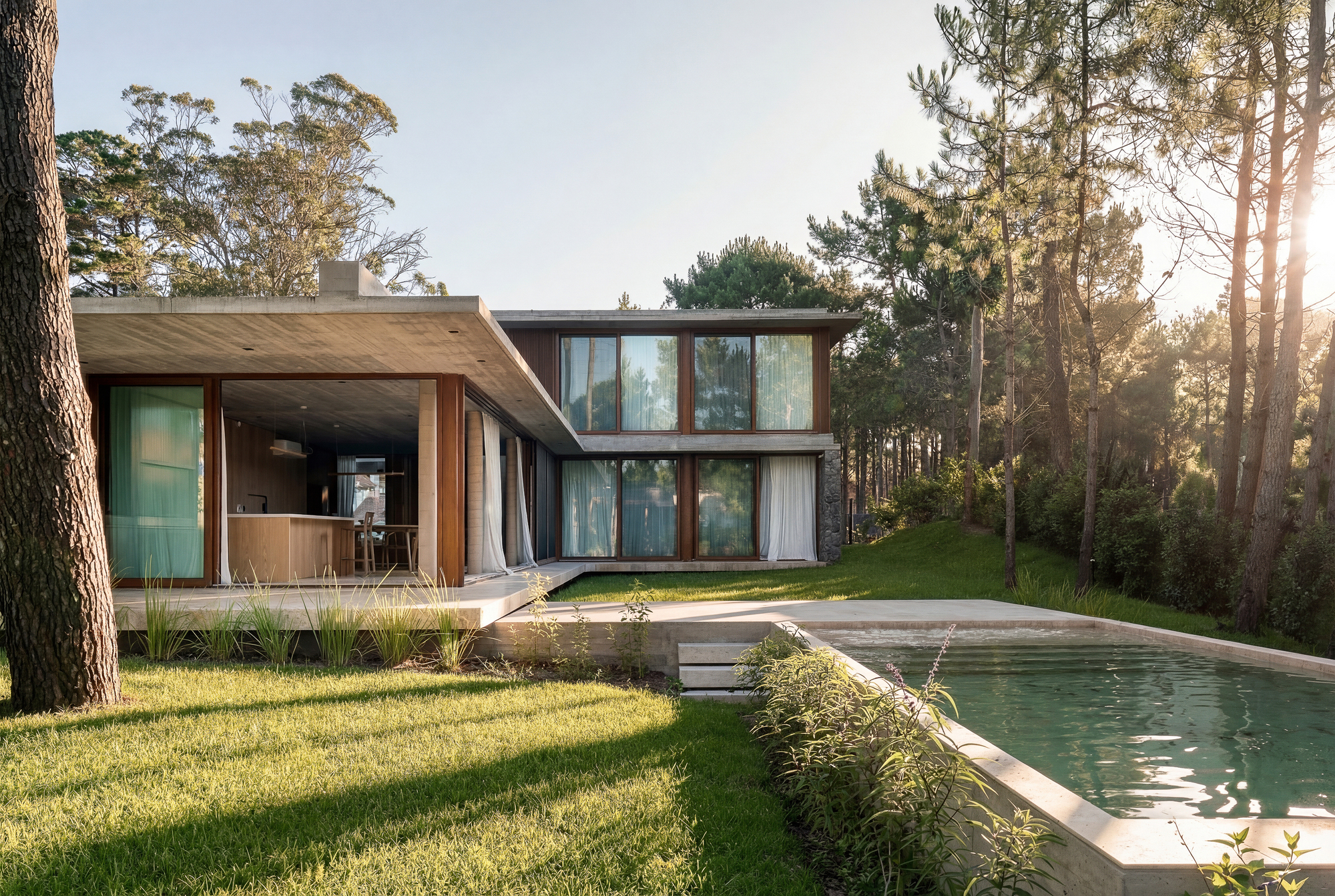 Two-storey house with a wide concrete roof overhang beside the pool, backlit by late sun through tall pines