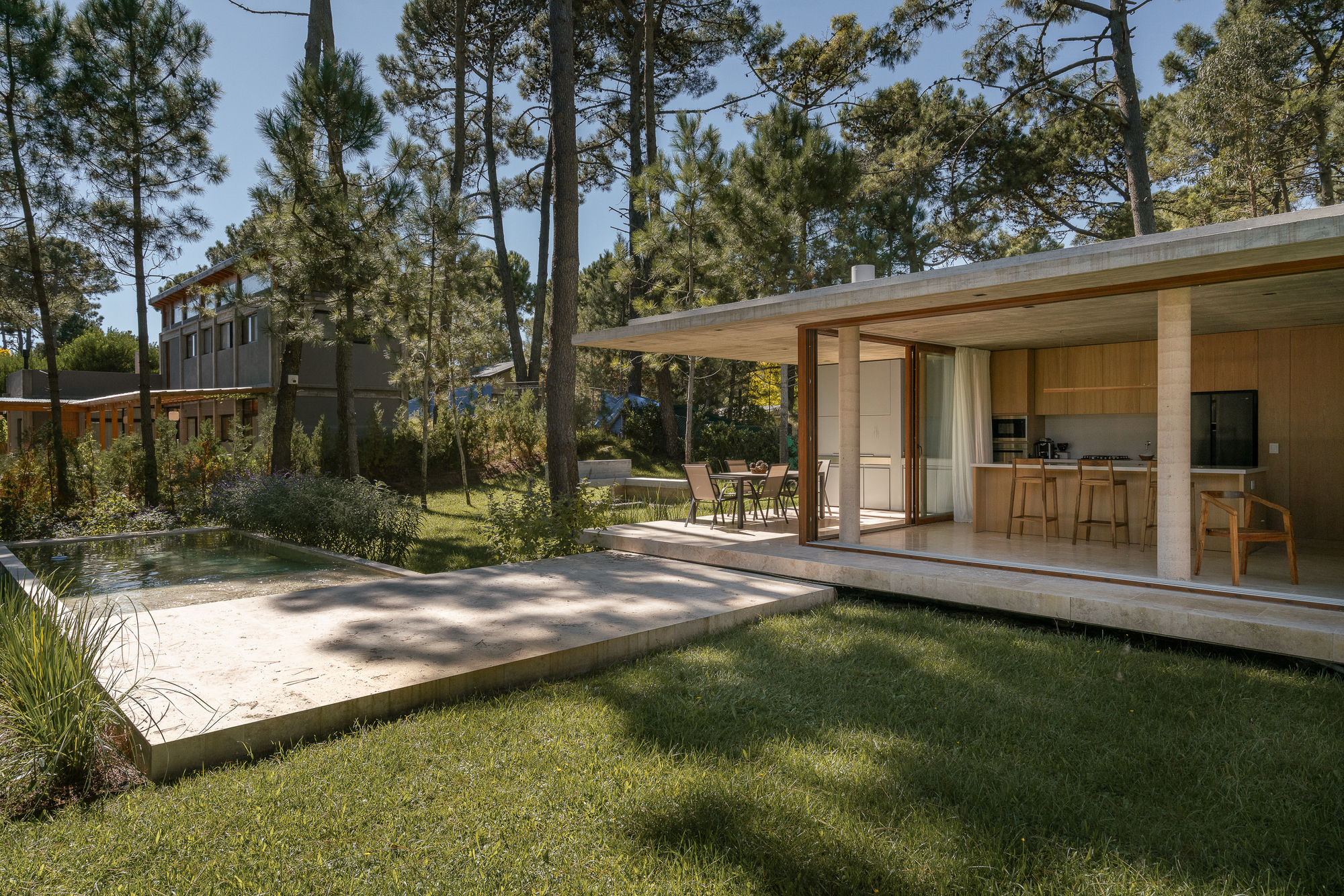 Flat-roofed concrete pavilion with open timber-framed glazing and kitchen bar, beside a stone-edged pool on a lawn dappled by pine shade