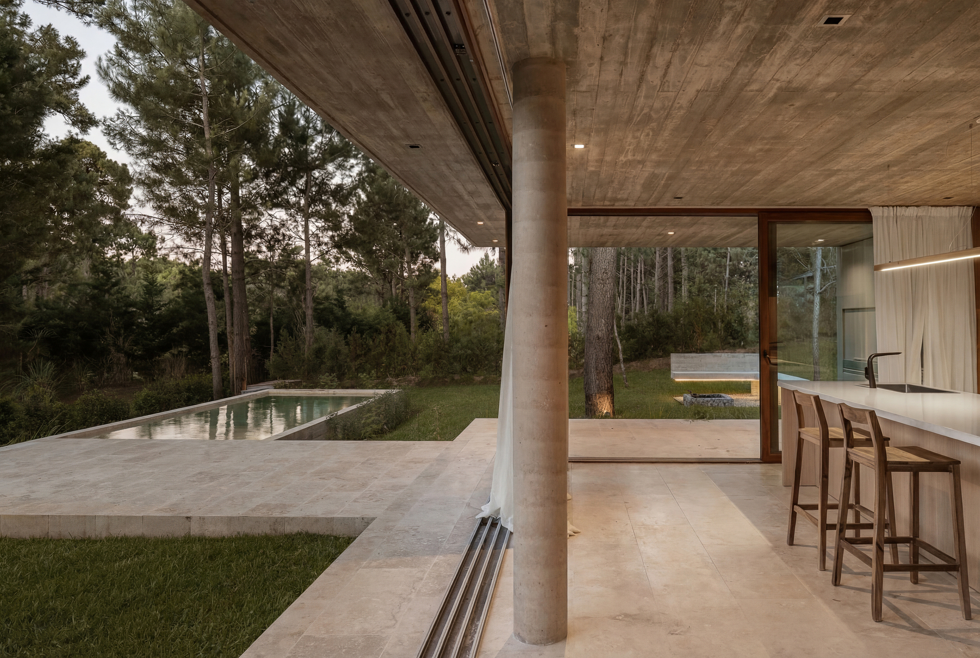 Board-formed concrete ceiling and round column over the kitchen bar with wooden stools, opening to the pool and pines at dusk