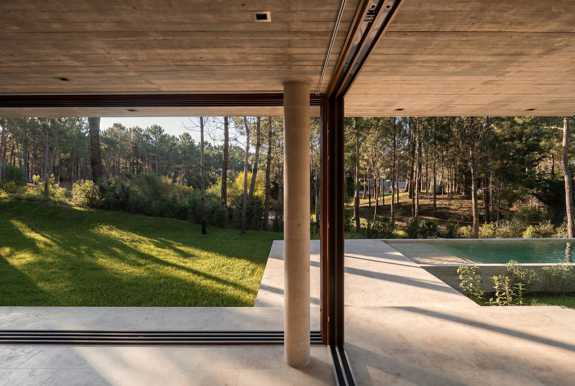 View from the covered terrace past a round concrete column and open sliding glass toward the pool and a sunlit lawn