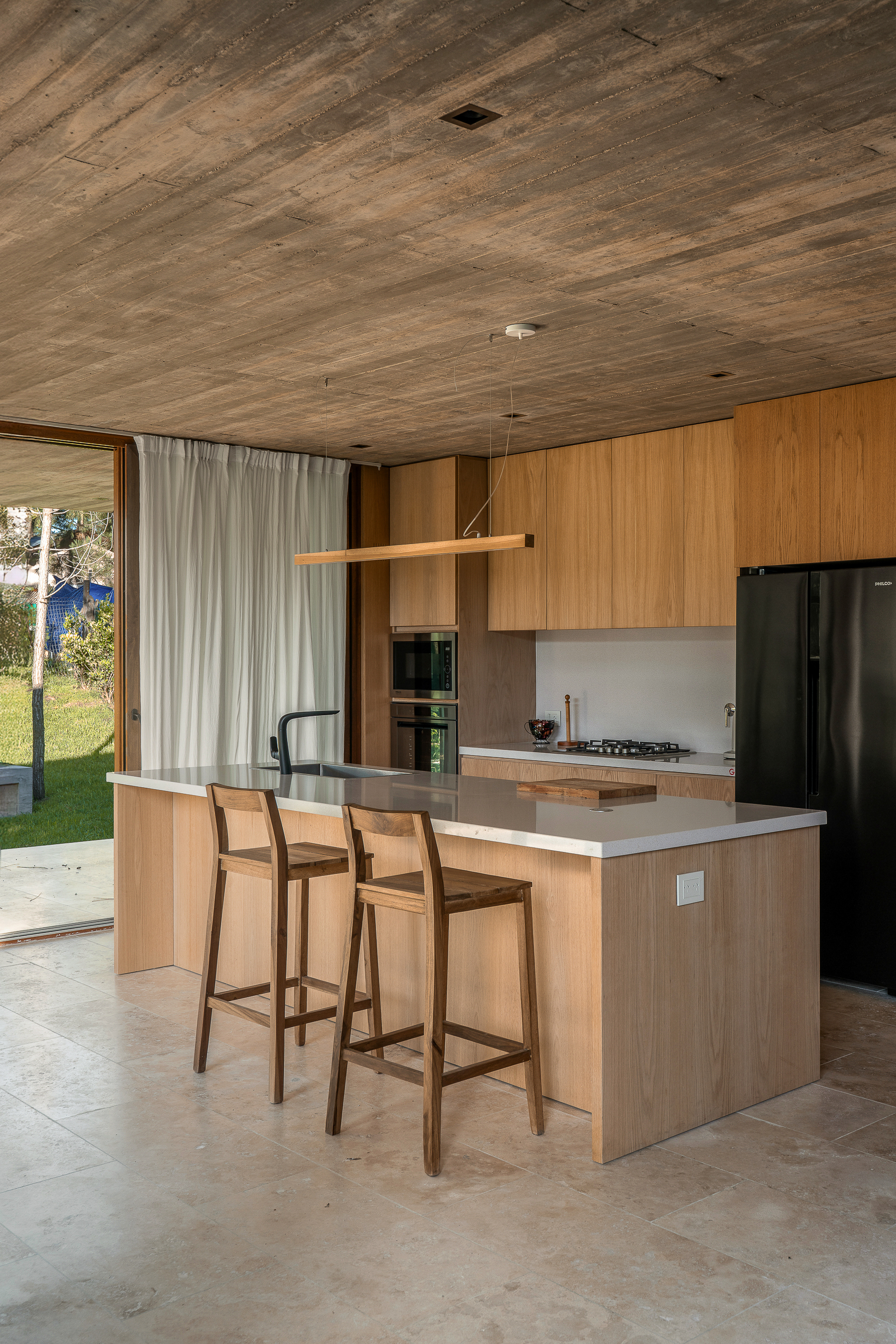 Kitchen island with two wooden stools, oak cabinetry and a linear pendant under a board-formed concrete ceiling in afternoon light