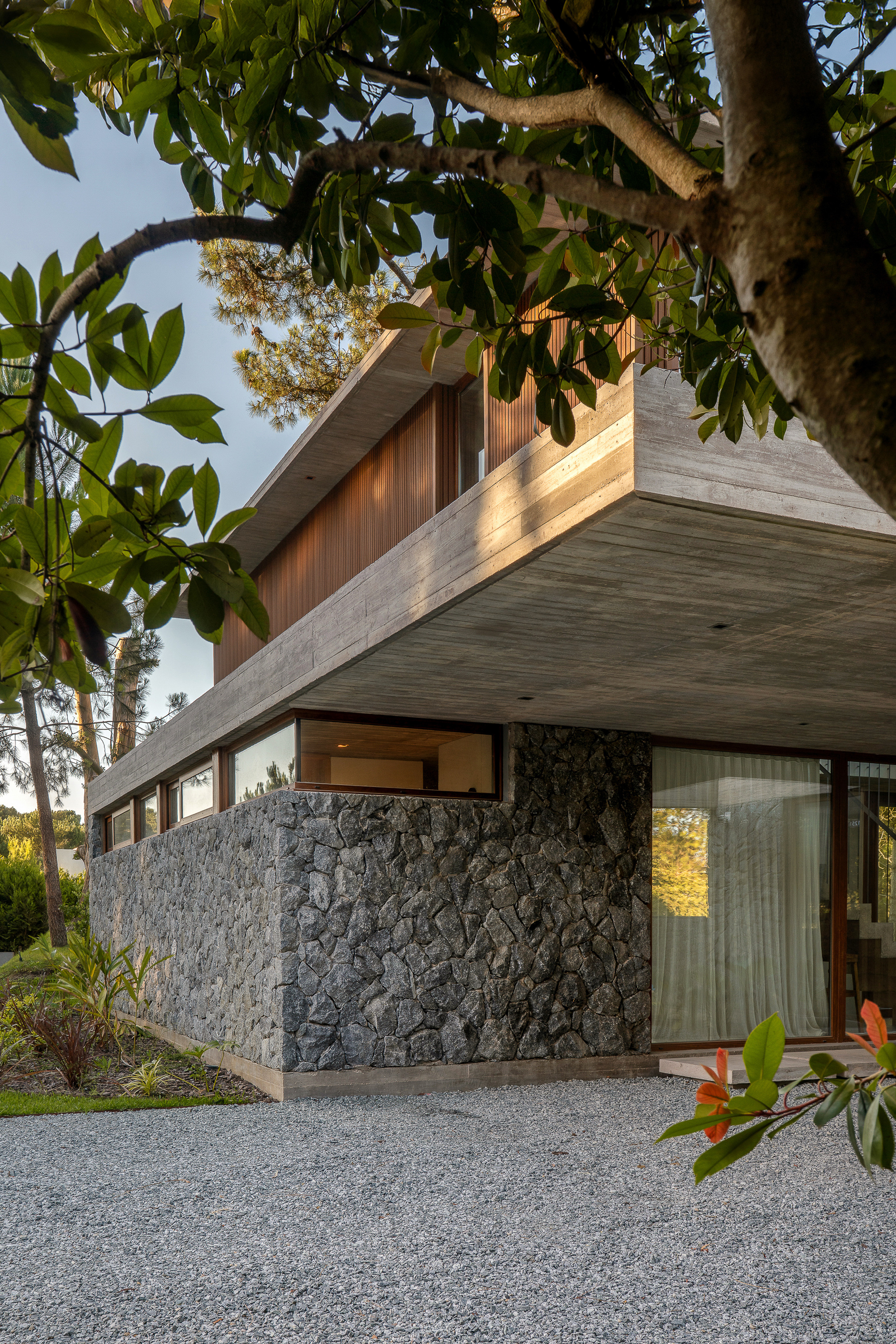 Corner of the upper volume cantilevering over a rough dark stone base, with a tree branch and gravel in the foreground