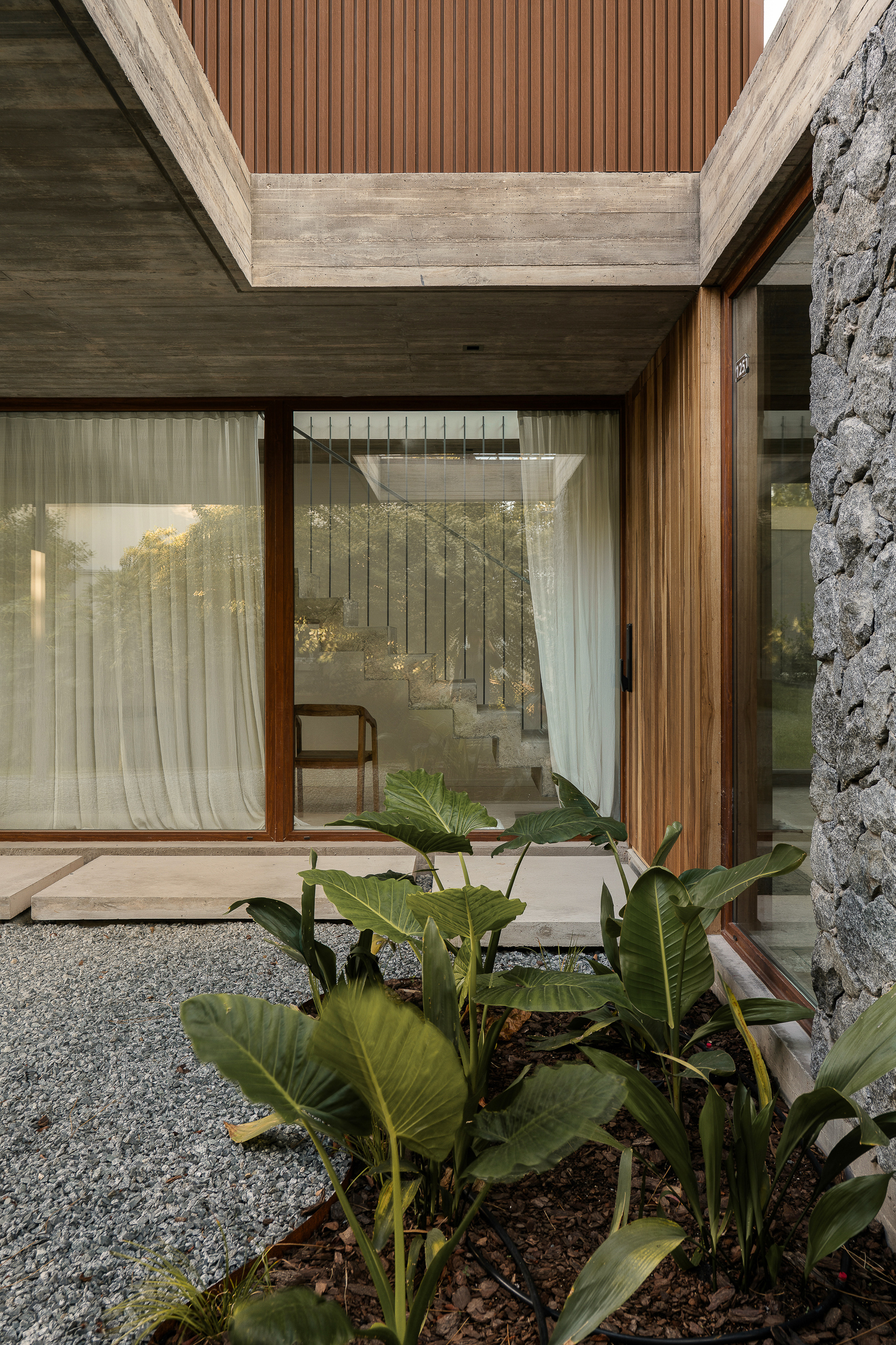 Entry court with broad-leaved plants and gravel in front of a glazed wall, timber slats and a stone edge, stair visible inside