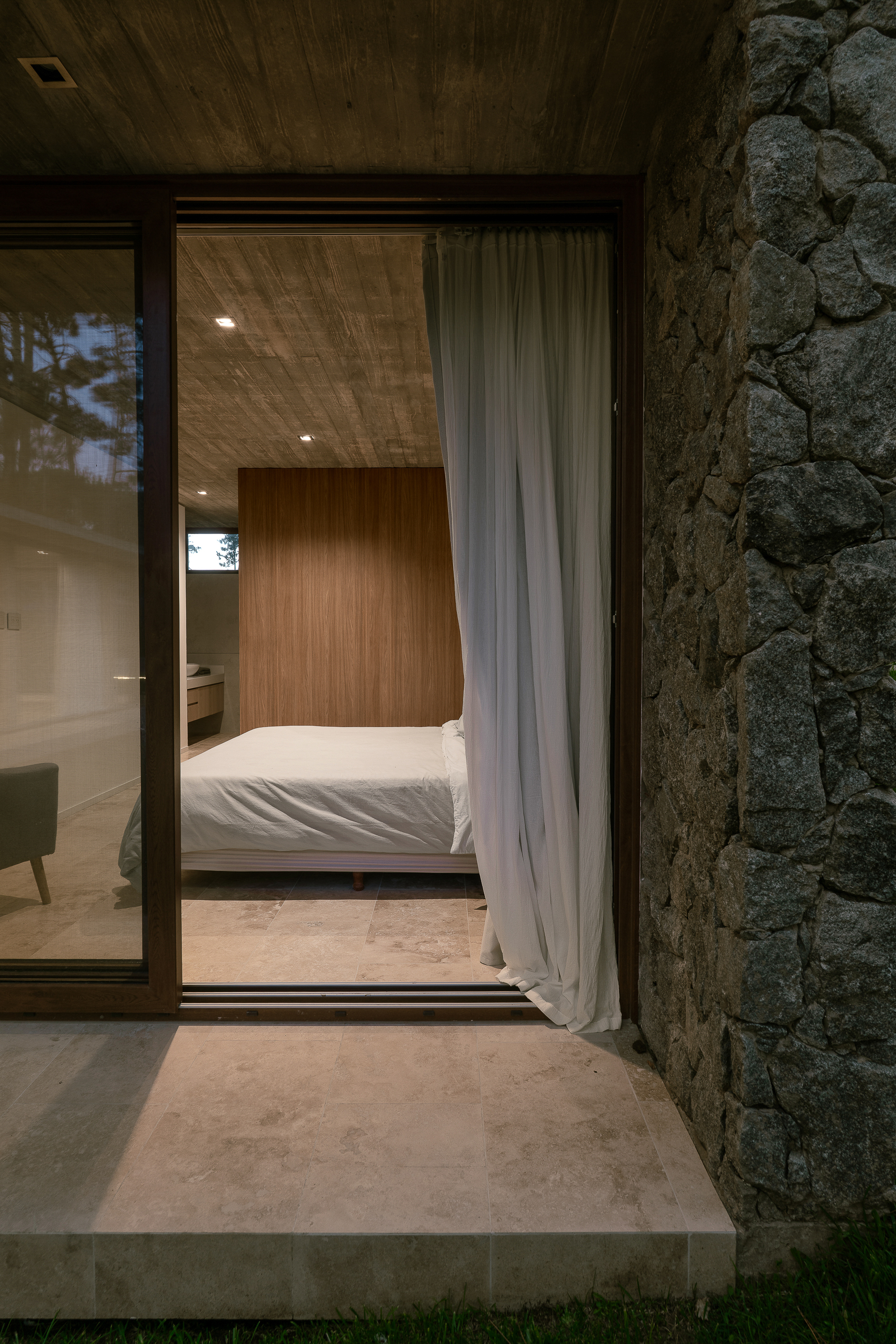 Bedroom seen through a sliding glass door with a sheer curtain, beside a rough fieldstone wall at dusk