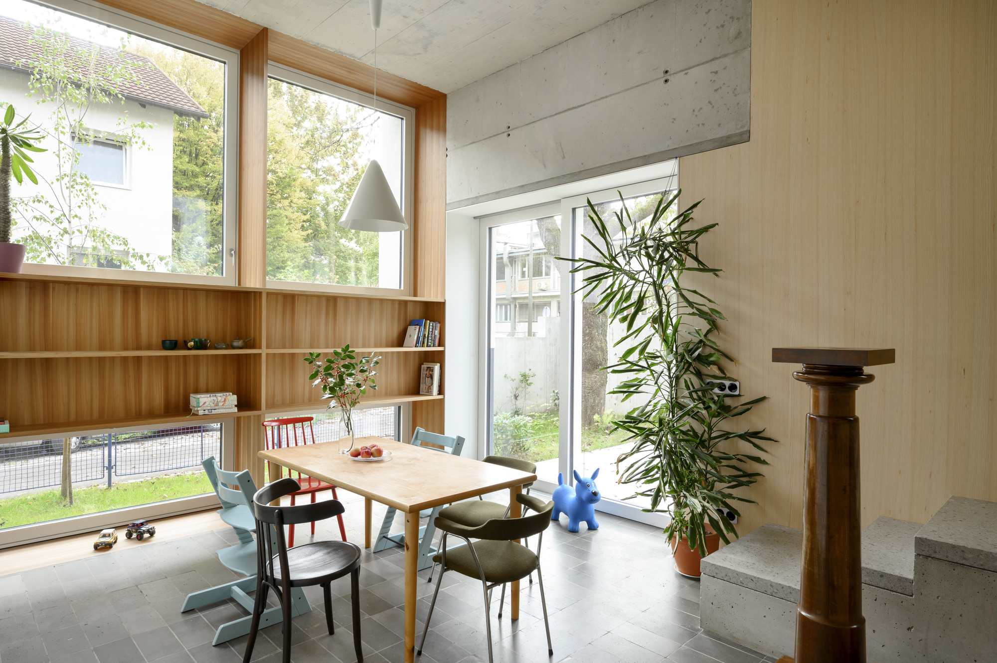 Dining area with timber shelving under tall windows, a concrete step and turned wooden column, lit by soft daylight from the garden