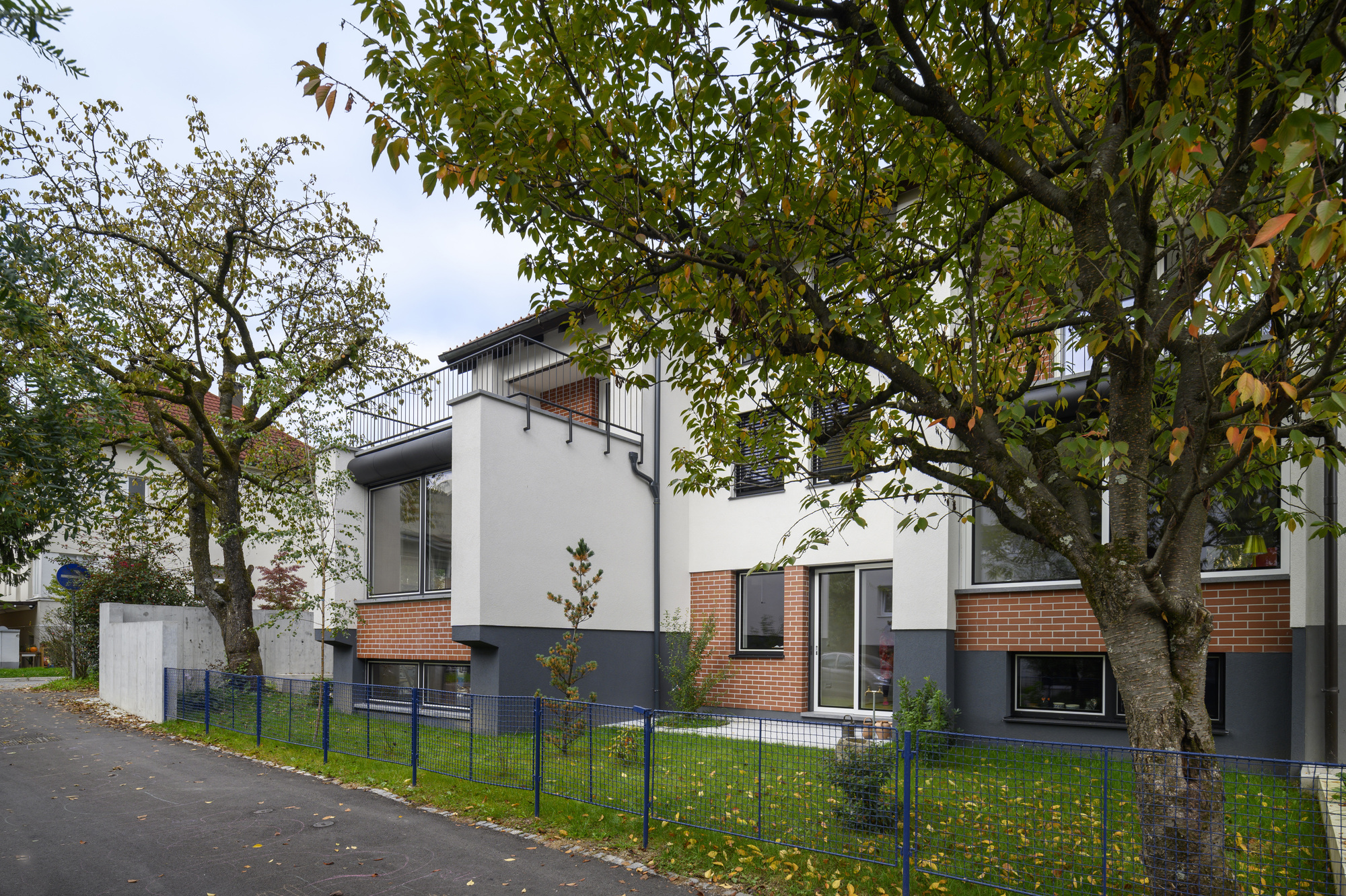 Street facade of white render and brick base behind a blue metal fence, framed by autumn cherry trees
