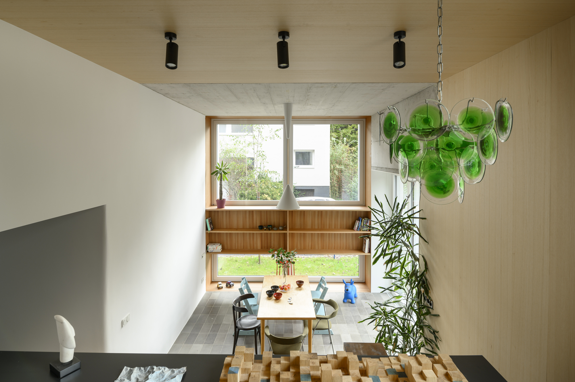 View down into a double-height dining space with green glass chandelier, timber ceiling and a wall of shelves beyond the table